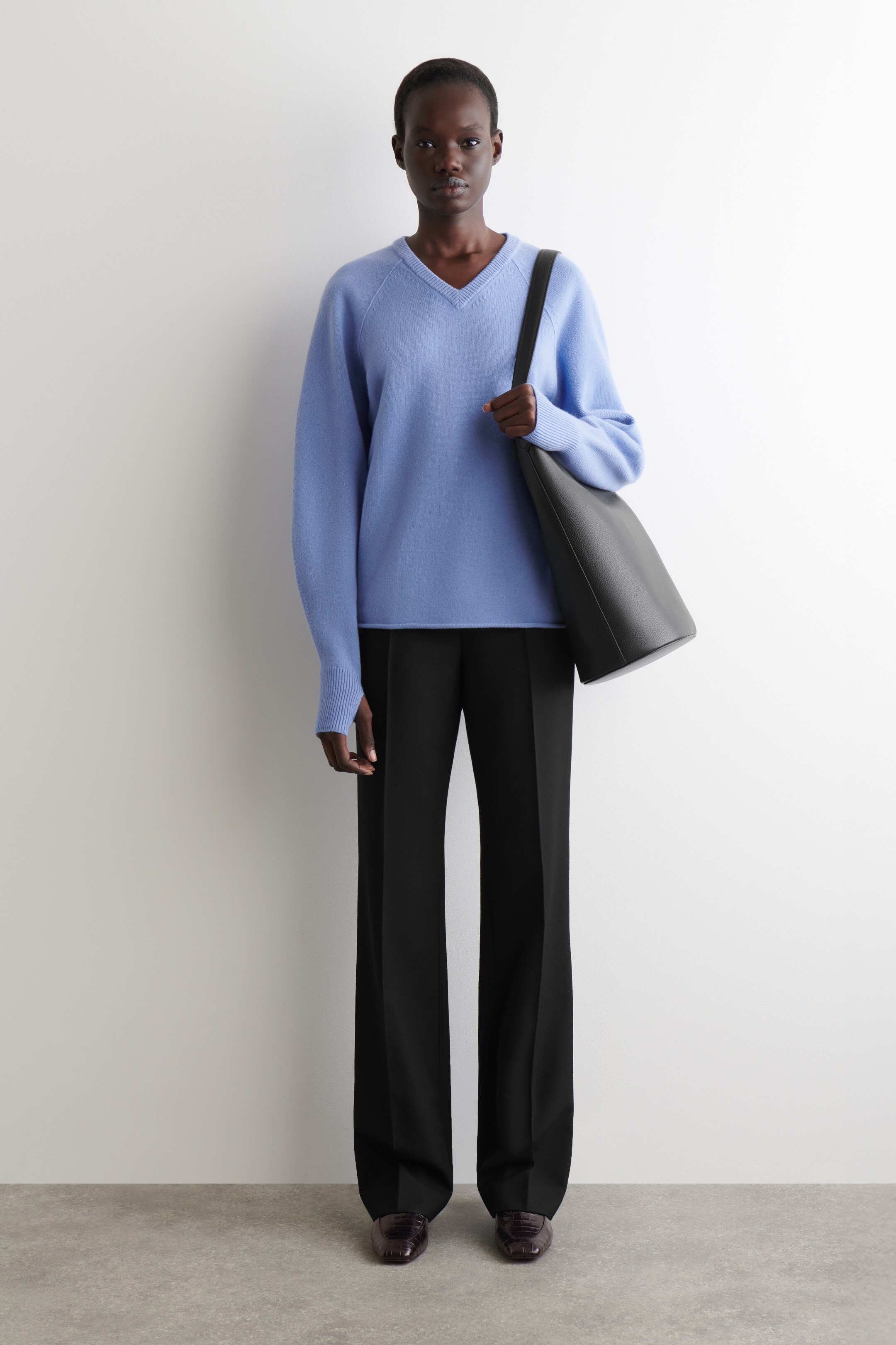 Ingrandisci l'immagine: Un maglione azzurro con scollo a V, caratterizzato da maniche raglan e un orlo a coste, viene indossato con pantaloni neri a gamba larga. Scarpe piatte marrone scuro e testurizzate completano il look, mentre una borsa tote nera e morbida viene portata sulla spalla sinistra.