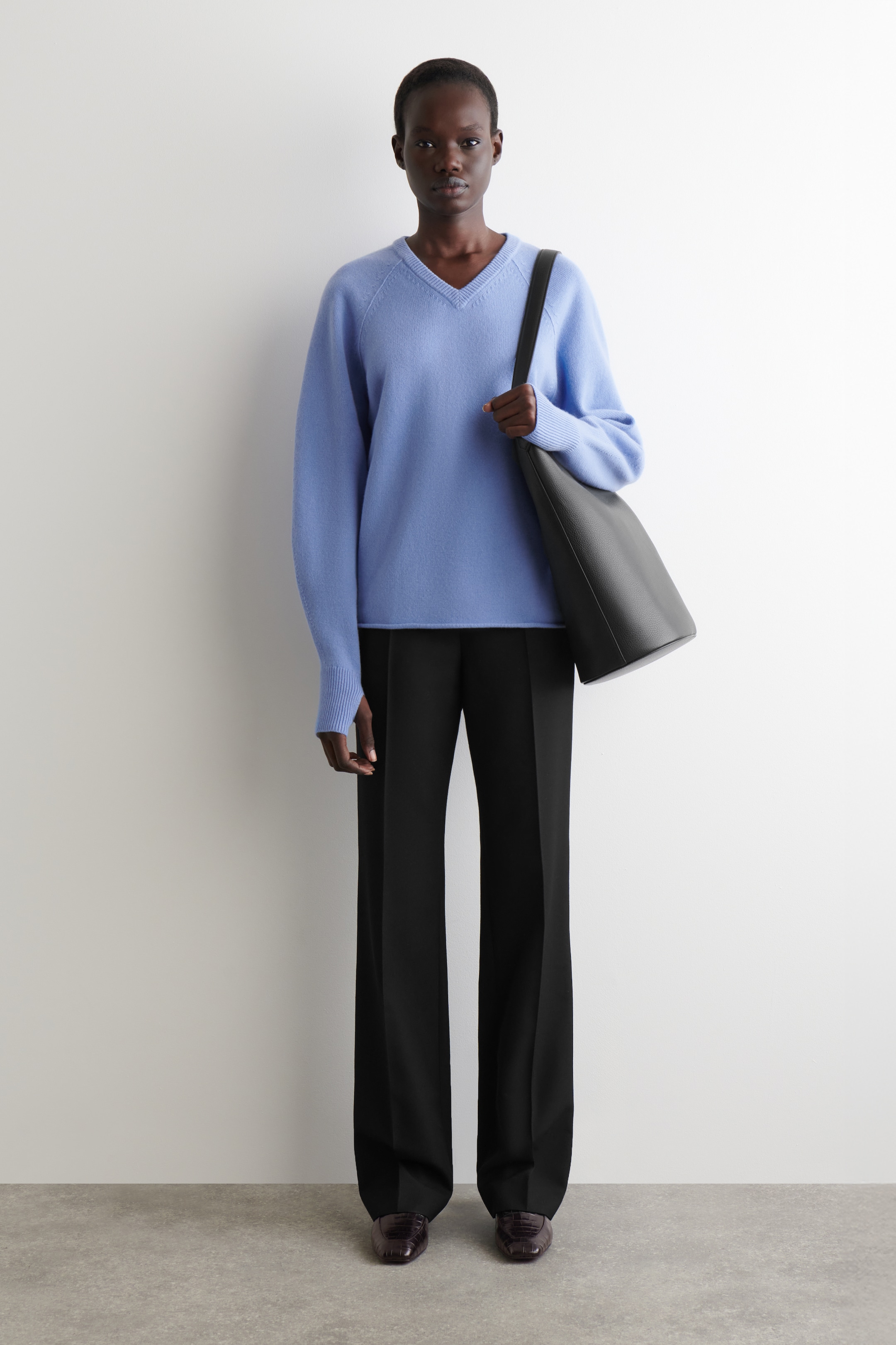 Visualizza immagine più grande: Un maglione azzurro con scollo a V, caratterizzato da maniche raglan e un orlo a coste, viene indossato con pantaloni neri a gamba larga. Scarpe piatte marrone scuro e testurizzate completano il look, mentre una borsa tote nera e morbida viene portata sulla spalla sinistra.