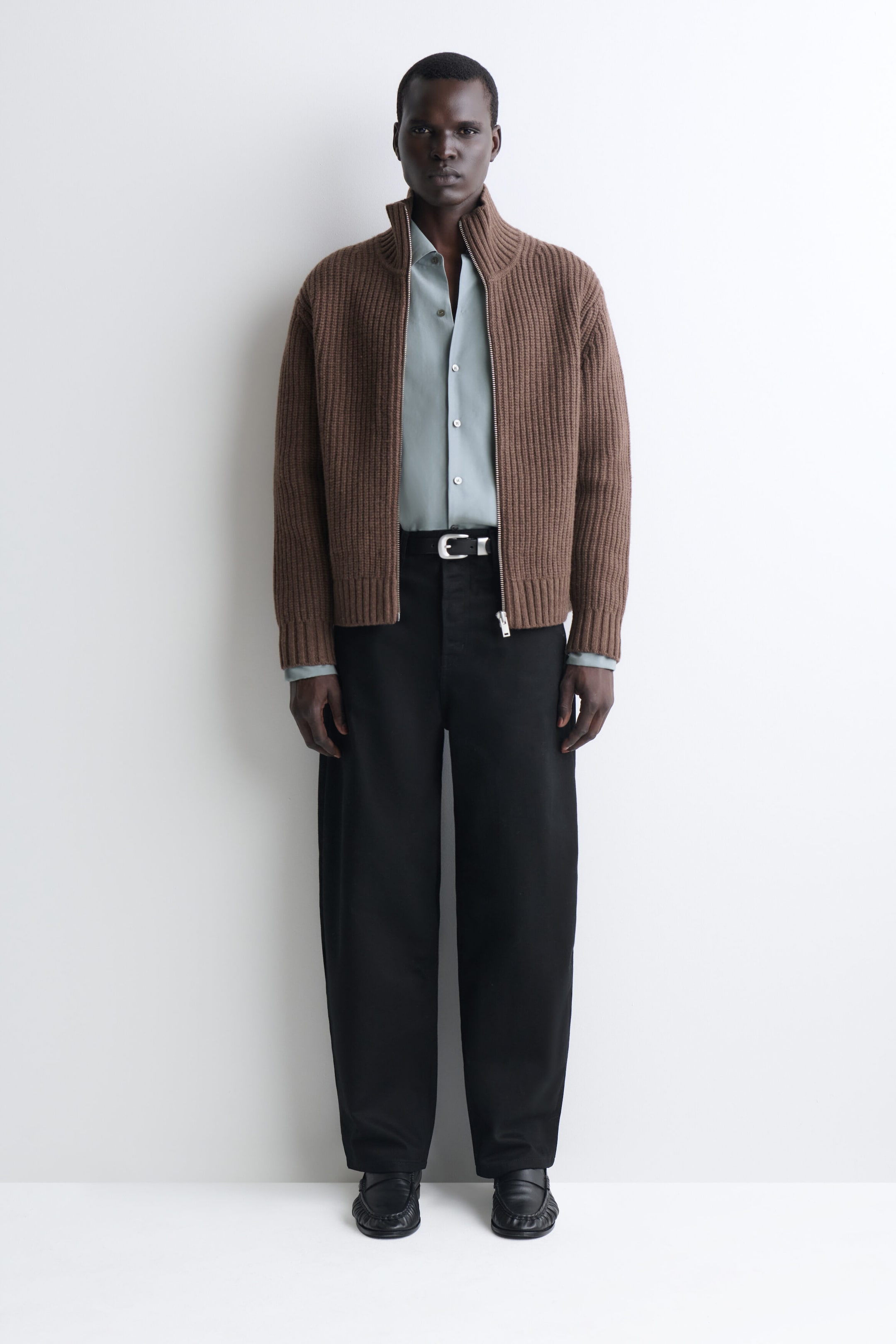 A brown ribbed-knit cardigan is shown from the front, featuring a high stand-up collar, long sleeves with ribbed cuffs, and a silver-tone front zipper. The cardigan is worn unzipped over a light-blue button-down shirt tucked into black trousers. A brown ribbed-knit cardigan is shown from the front, featuring a high stand-up collar, long sleeves with ribbed cuffs, and a silver-tone front zipper. The cardigan is worn unzipped over a light-blue button-down shirt tucked into black trousers.