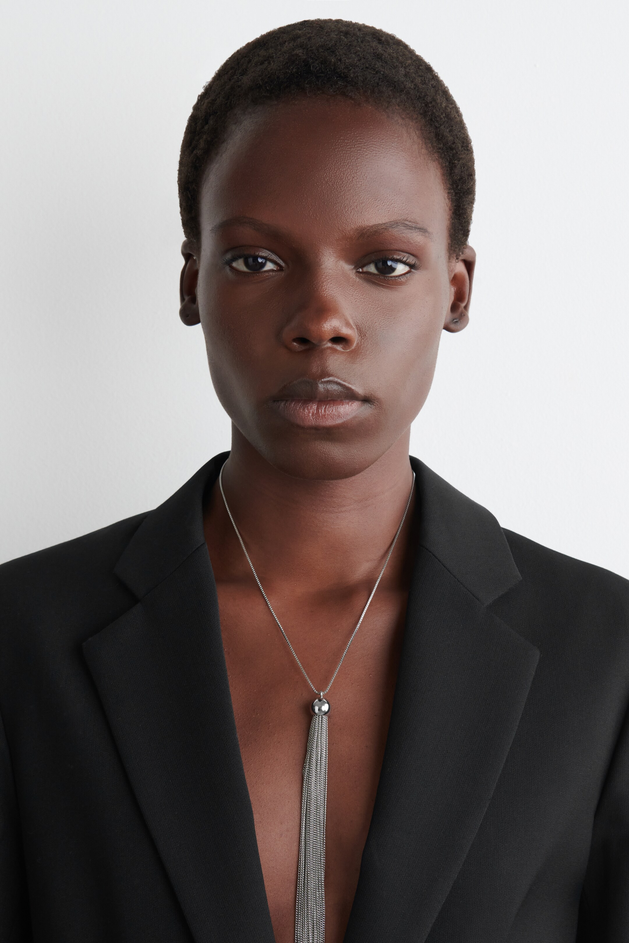 Ver imagen más grande: Un collar de tono plateado cuelga centralmente en el pecho, con un colgante esférico del que penden múltiples cadenas finas, formando un fleco. Descansa contra las solapas abiertas de un blazer negro.