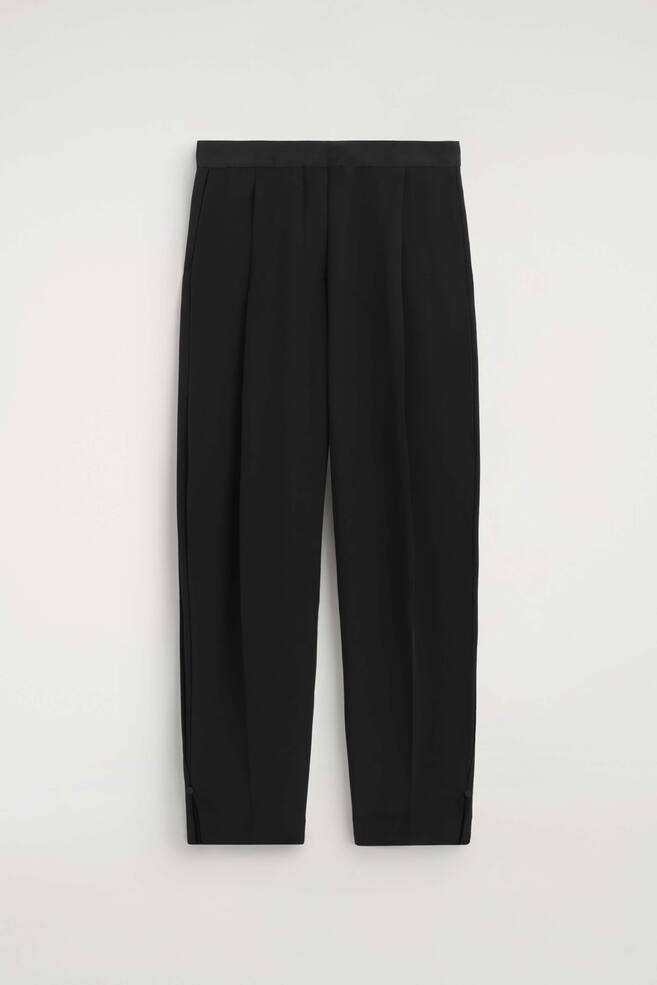 Black trousers are shown facing the front, featuring a structured waistband and prominent front pleats that taper down the legs. The trousers are presented in a relaxed fit with small slit details and button fastenings at the outer ankle hems. - 1