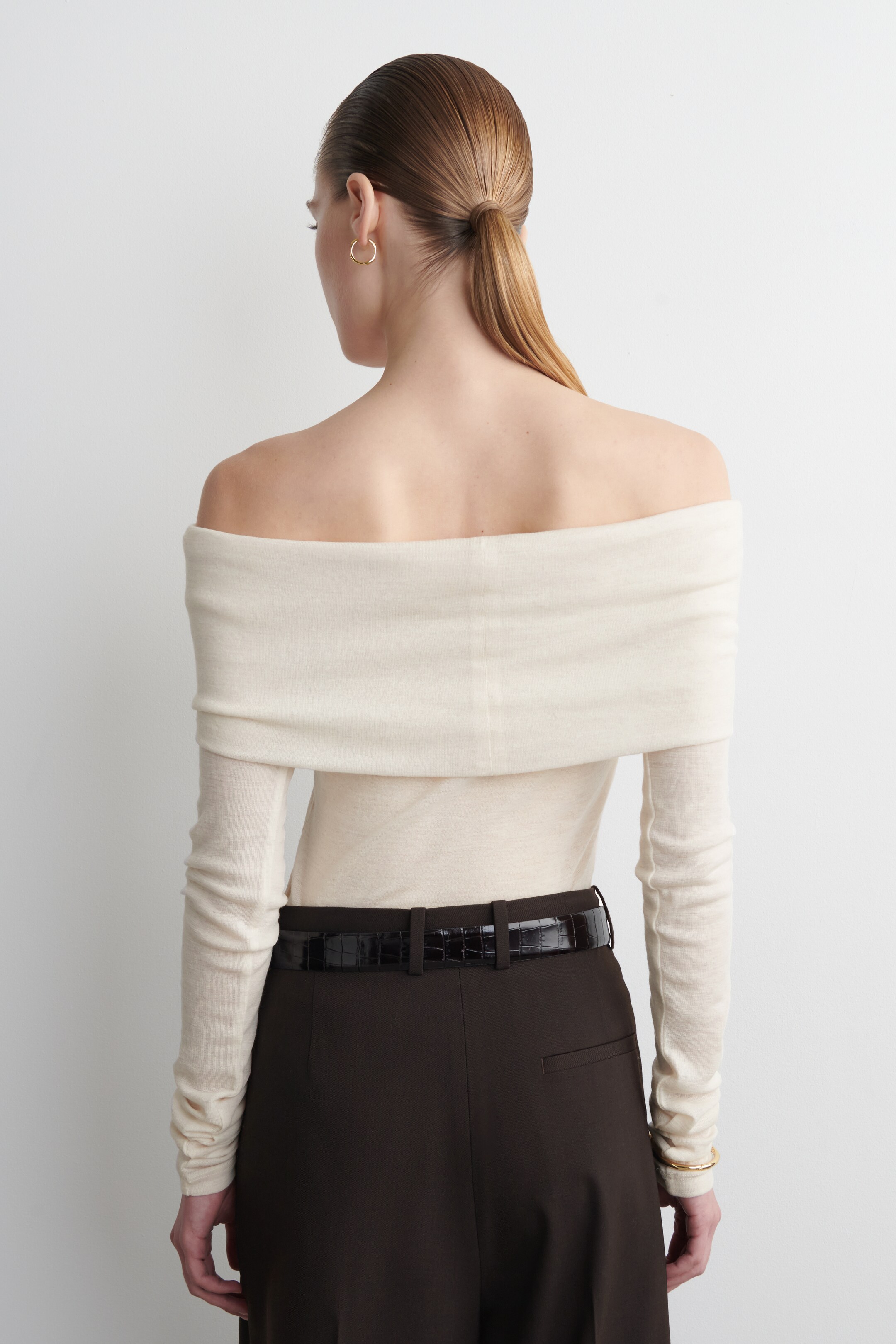 Agrandir l'image: Un haut crème à épaules dénudées est vu de dos, avec un large décolleté drapé s'étendant sur les épaules et de longues manches. Il est rentré dans un pantalon brun foncé, avec une ceinture en cuir embossé de motif crocodile visible autour de la taille.