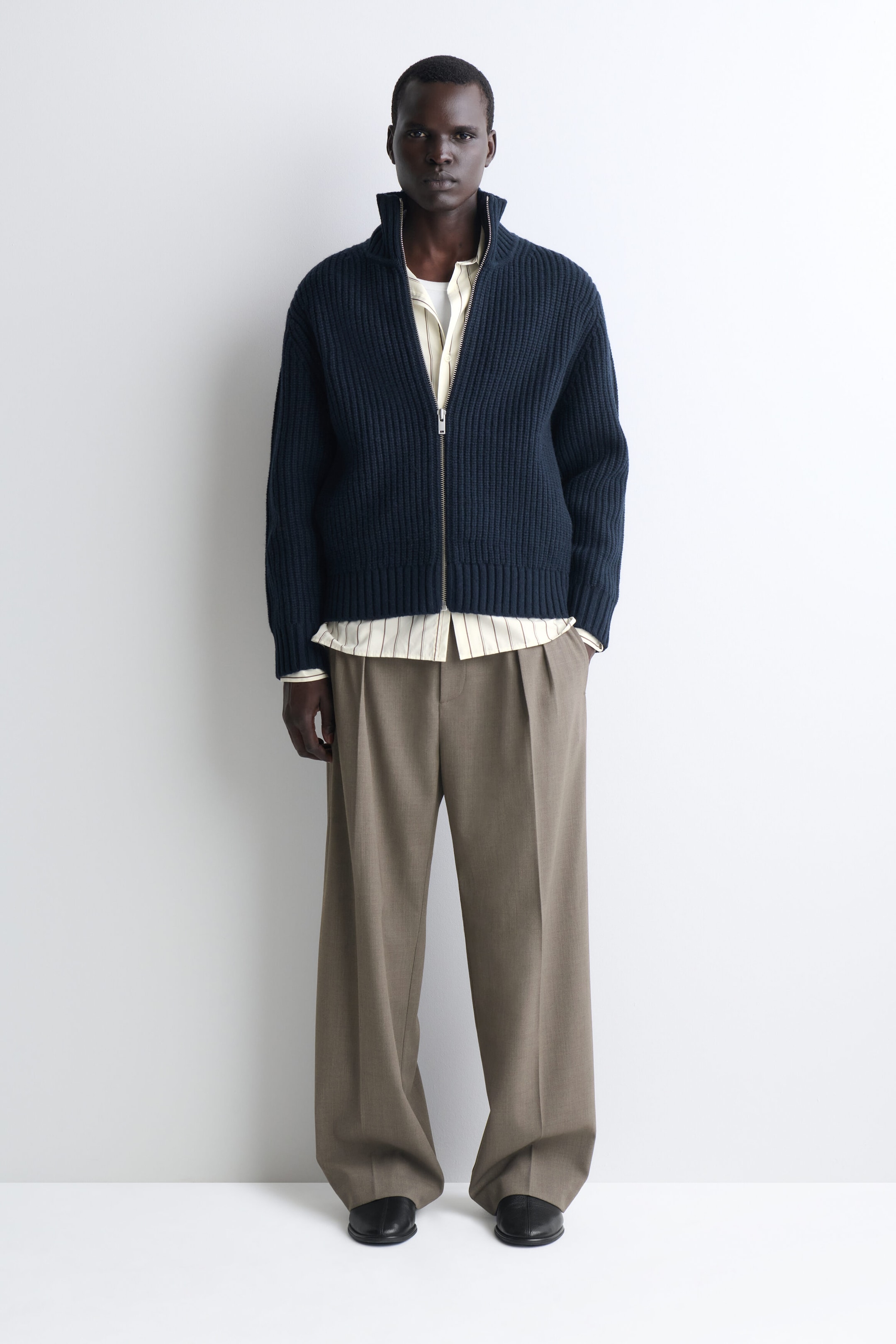 This front-facing view highlights a navy blue, heavyweight rib-knit zip-up cardigan featuring a high stand-up collar and a silver front zipper. The cardigan is layered over a cream pinstriped shirt and paired with taupe wide-leg pleated trousers. This front-facing view highlights a navy blue, heavyweight rib-knit zip-up cardigan featuring a high stand-up collar and a silver front zipper. The cardigan is layered over a cream pinstriped shirt and paired with taupe wide-leg pleated trousers.