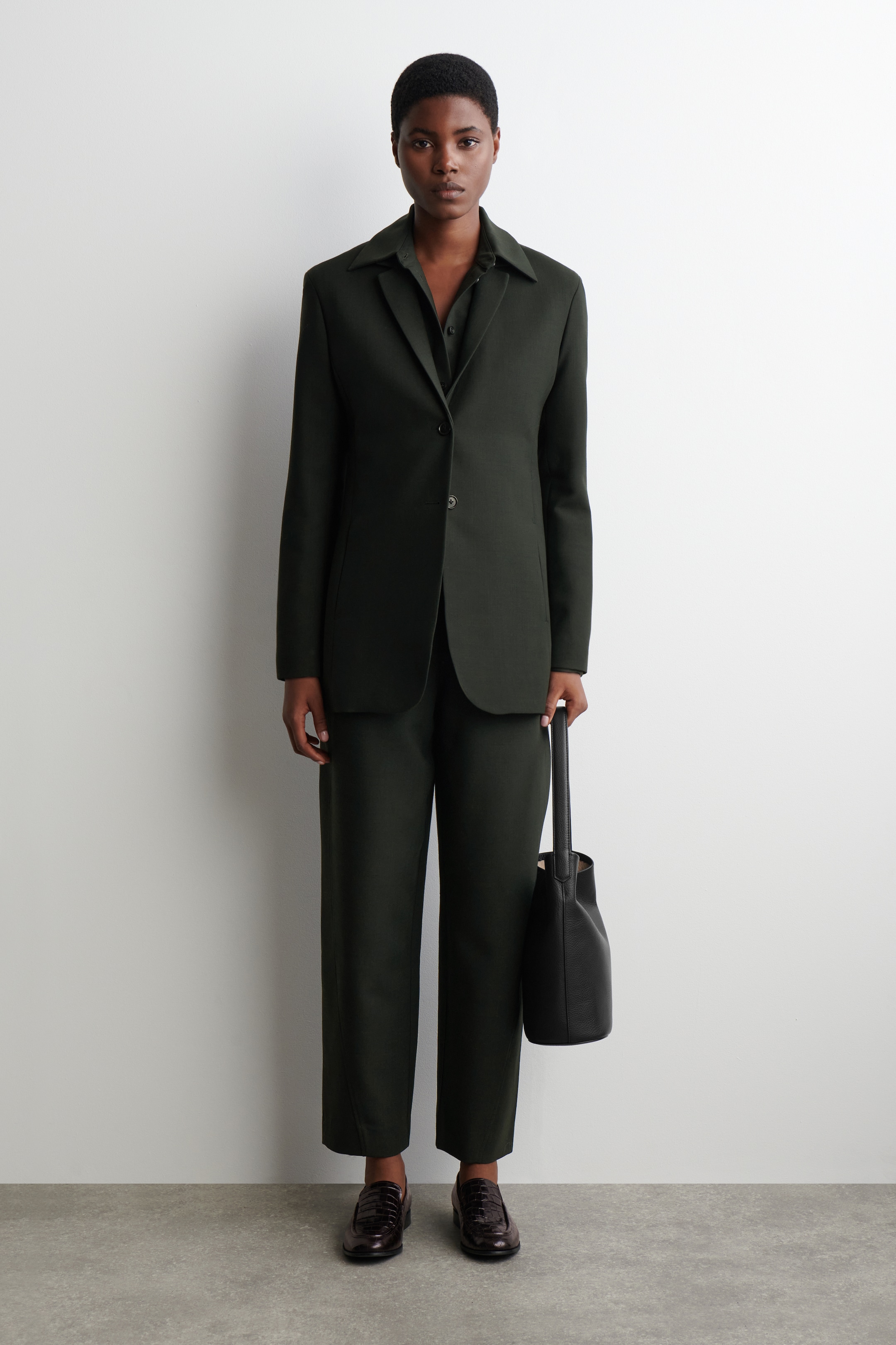 Grösseres Bild ansehen: Ein dunkelgrüner, einreihiger Blazer mit einem gespreizten Hemdkragen und einem Ein-Knopf-Verschluss wird frontseitig über einem passenden Hemd mit Kragen getragen und mit locker sitzenden Hosen kombiniert. Eine schwarze Leder-Bucket-Bag wird gehalten, und dunkelbraune, strukturierte Loafer werden getragen.