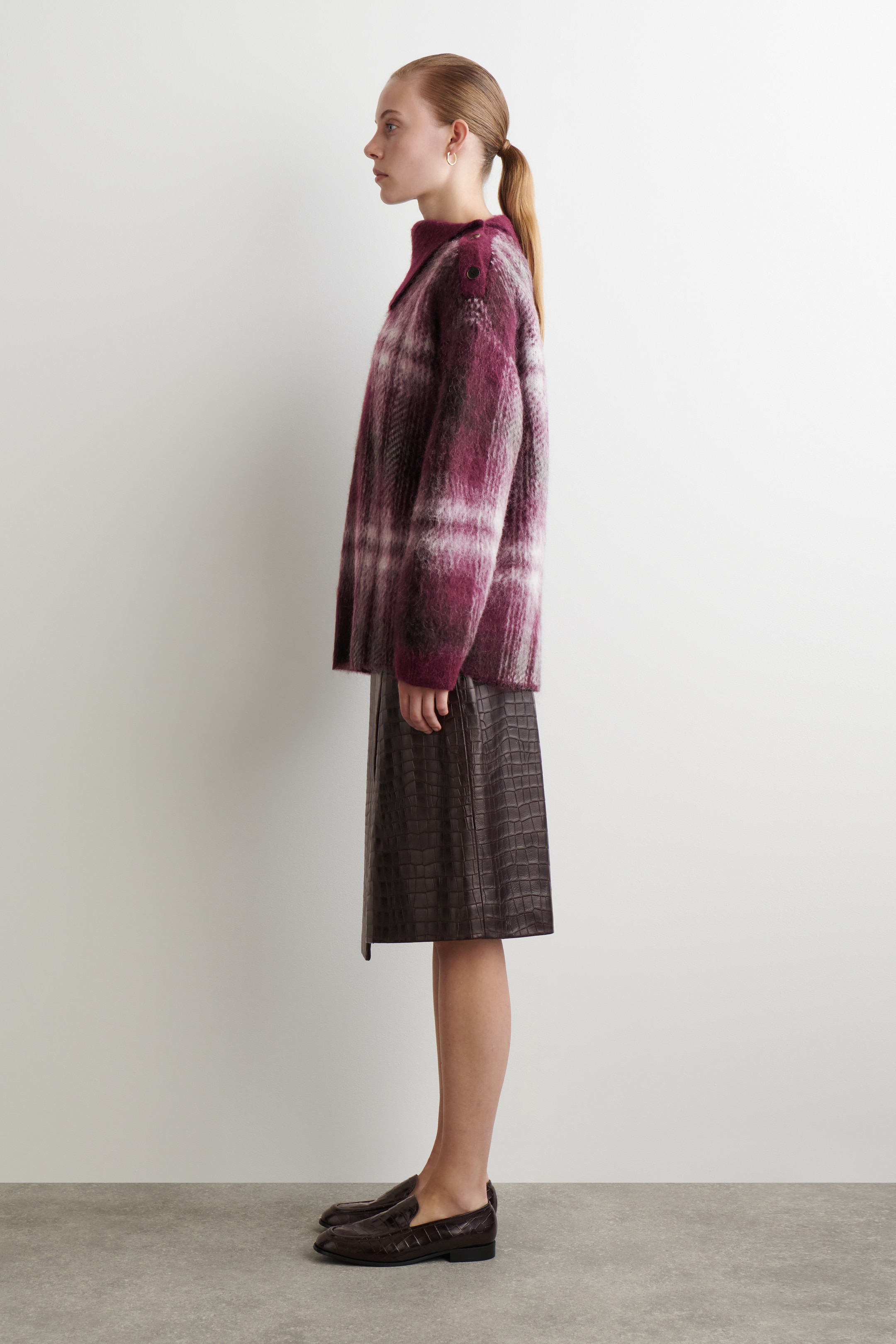 Agrandir l'image: Un pull en plaid duveteux dans des tons de violet et de blanc est présenté de profil. Il présente une coupe ample, un col montant et deux boutons sombres sur chaque épaule, et est porté sur une jupe marron foncé à motif crocodile.