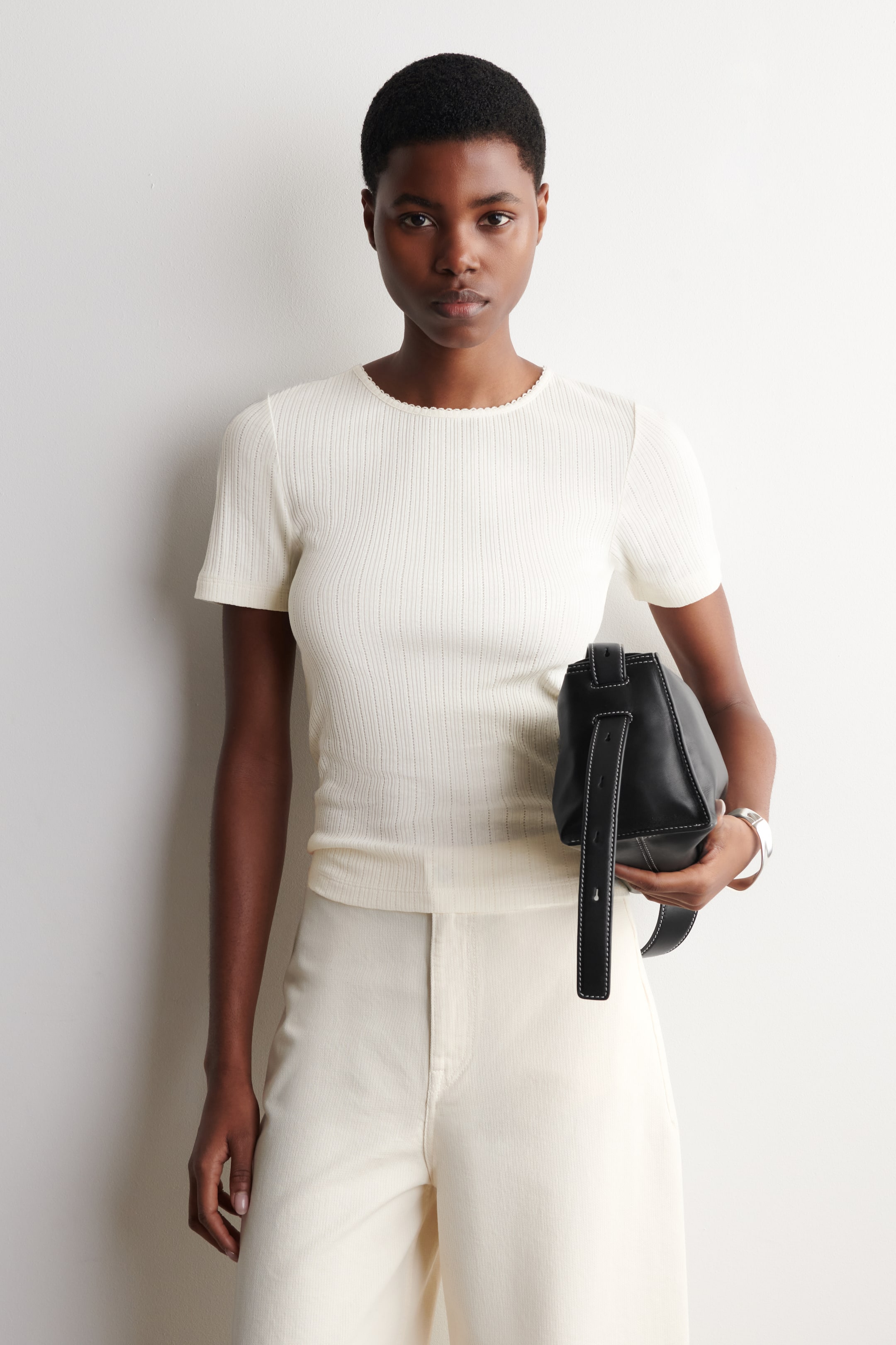 Größeres Bild ansehen: Ein cremeweißes, figurbetontes Kurzarm-Top mit einem gewellten Rundhalsausschnitt und feiner vertikaler Rippung wird getragen, eingesteckt in cremefarbene, hoch taillierte Hosen mit geradem Bein.