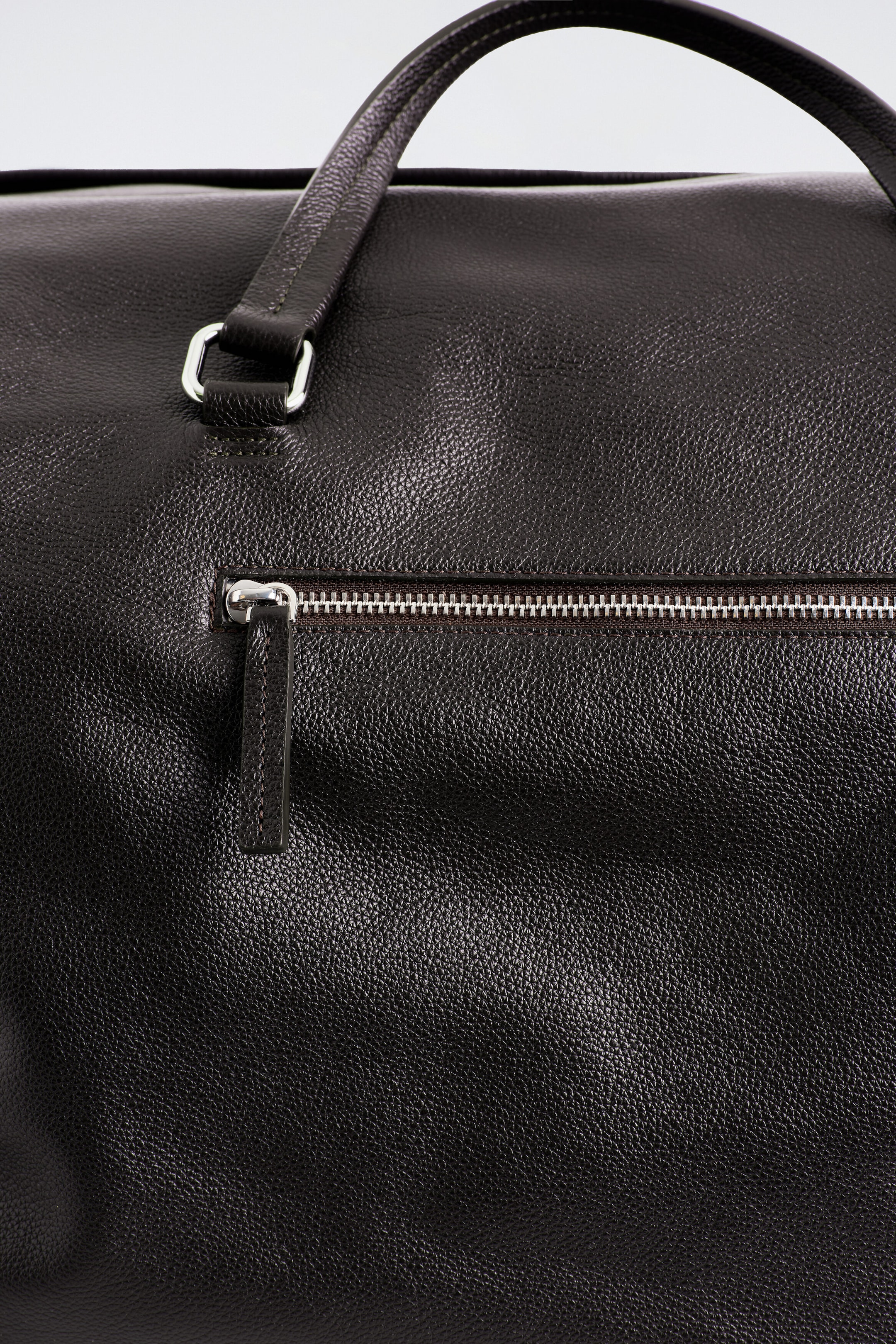 The Departure Duffle Bag - Leather is showcasing its dark brown pebbled leather texture in a close-up front view. The bag features a horizontal front pocket with a silver-tone zip and a leather pull tab, alongside a rolled leather top handle attached by a silver-tone metal ring.