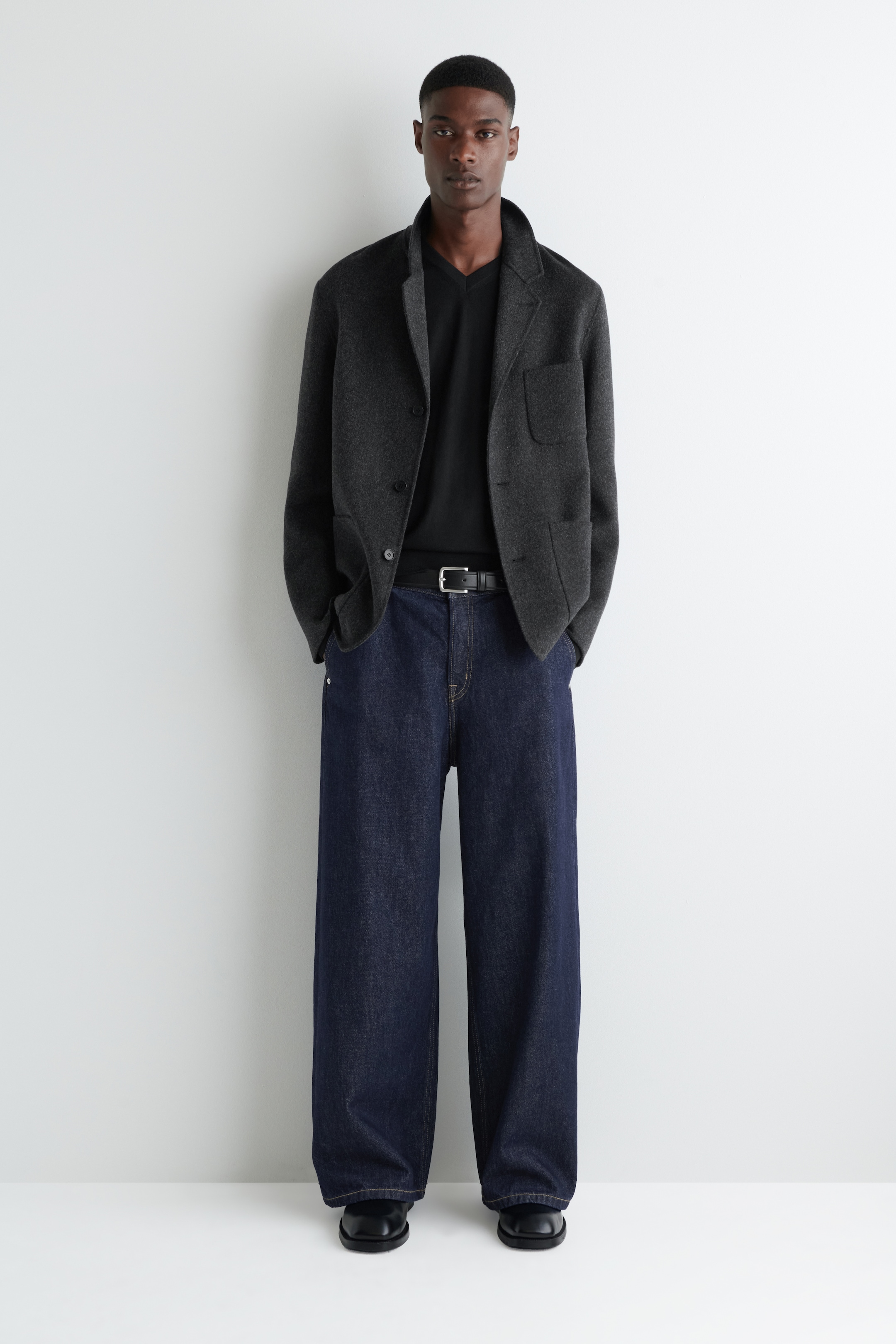 Agrandir l'image: Un ensemble de vêtements pour hommes orienté vers l'avant est présenté. Un blazer gris charbon à deux boutons, avec de larges revers crantés et des poches plaquées, est superposé à un t-shirt noir à col en V. Un jean indigo foncé à jambes larges est porté, maintenu par une ceinture en cuir noir et associé à des chaussures noires à bout carré.
