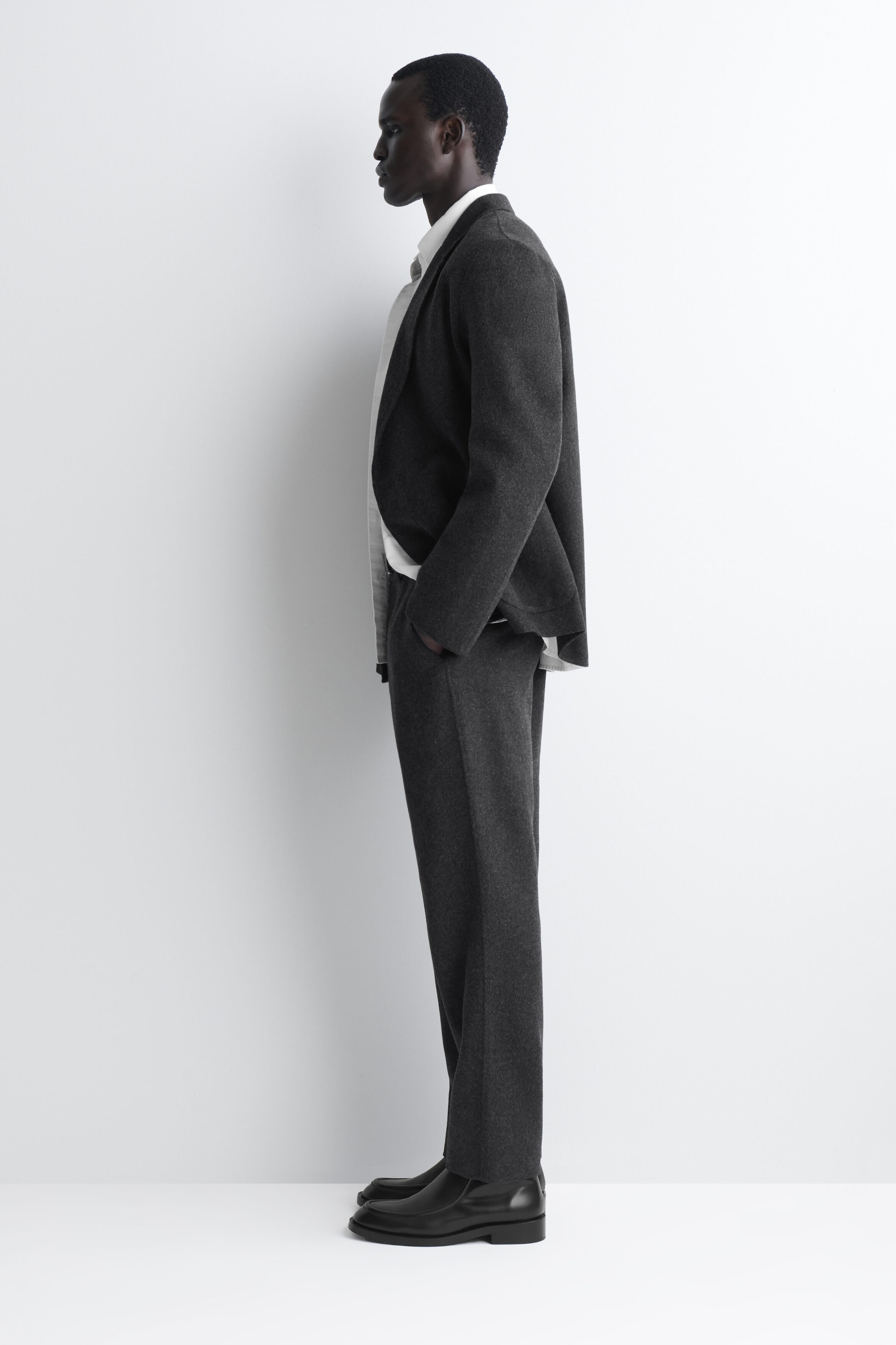 These dark grey wool-blend trousers, shown facing left in a side-profile view, are draping in an unstructured, relaxed straight-leg shape. They are being worn with a matching dark grey blazer, a white collared shirt, and black leather shoes to showcase their soft texture and clean silhouette. These dark grey wool-blend trousers, shown facing left in a side-profile view, are draping in an unstructured, relaxed straight-leg shape. They are being worn with a matching dark grey blazer, a white collared shirt, and black leather shoes to showcase their soft texture and clean silhouette.
