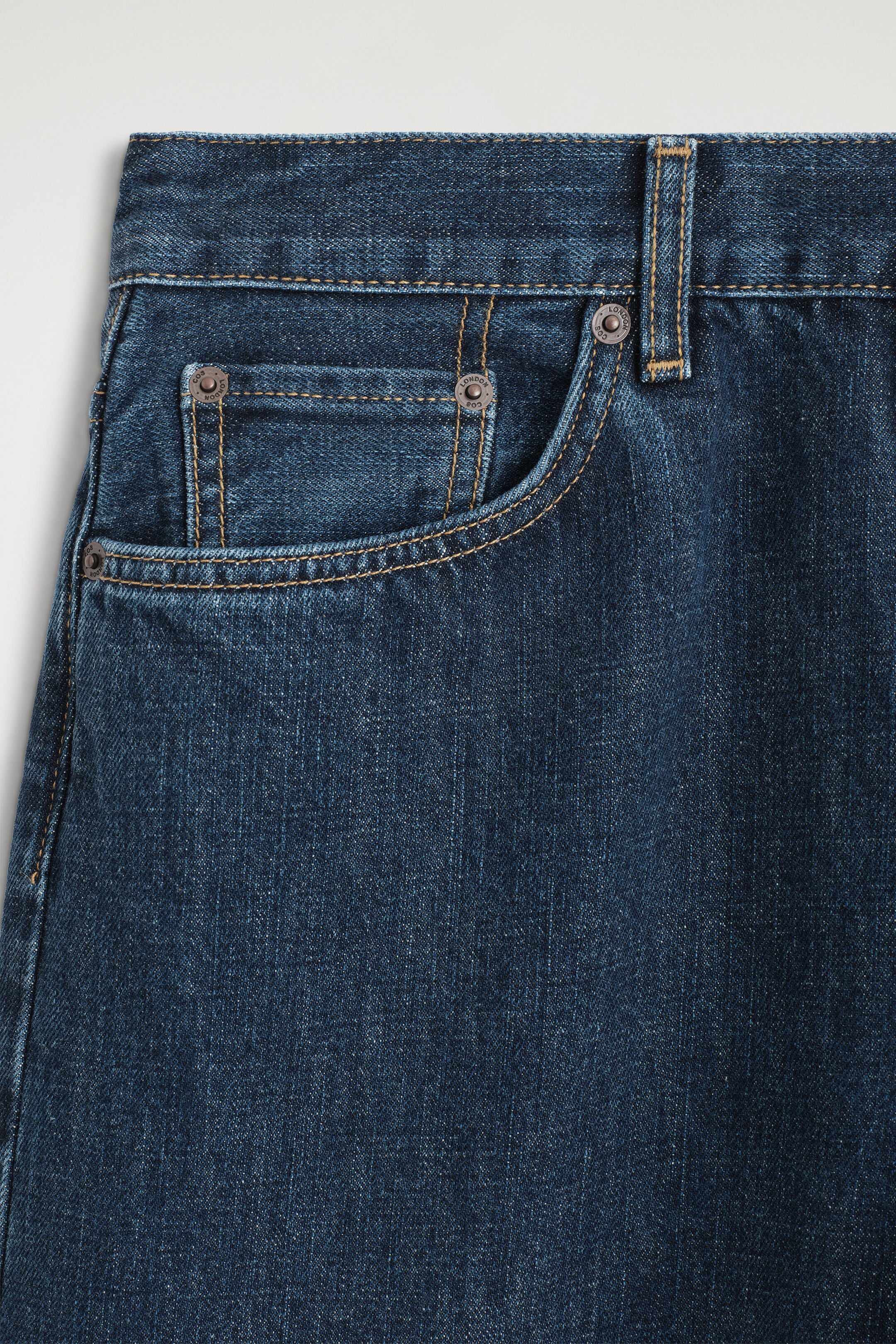 Dark blue denim jeans are shown from the upper right front, featuring a main curved pocket and a small coin pocket. Both pockets include metal rivets, some engraved with "LONDON COS," and are accented with contrasting orange stitching along the seams and waistband. Dark blue denim jeans are shown from the upper right front, featuring a main curved pocket and a small coin pocket. Both pockets include metal rivets, some engraved with "LONDON COS," and are accented with contrasting orange stitching along the seams and waistband.