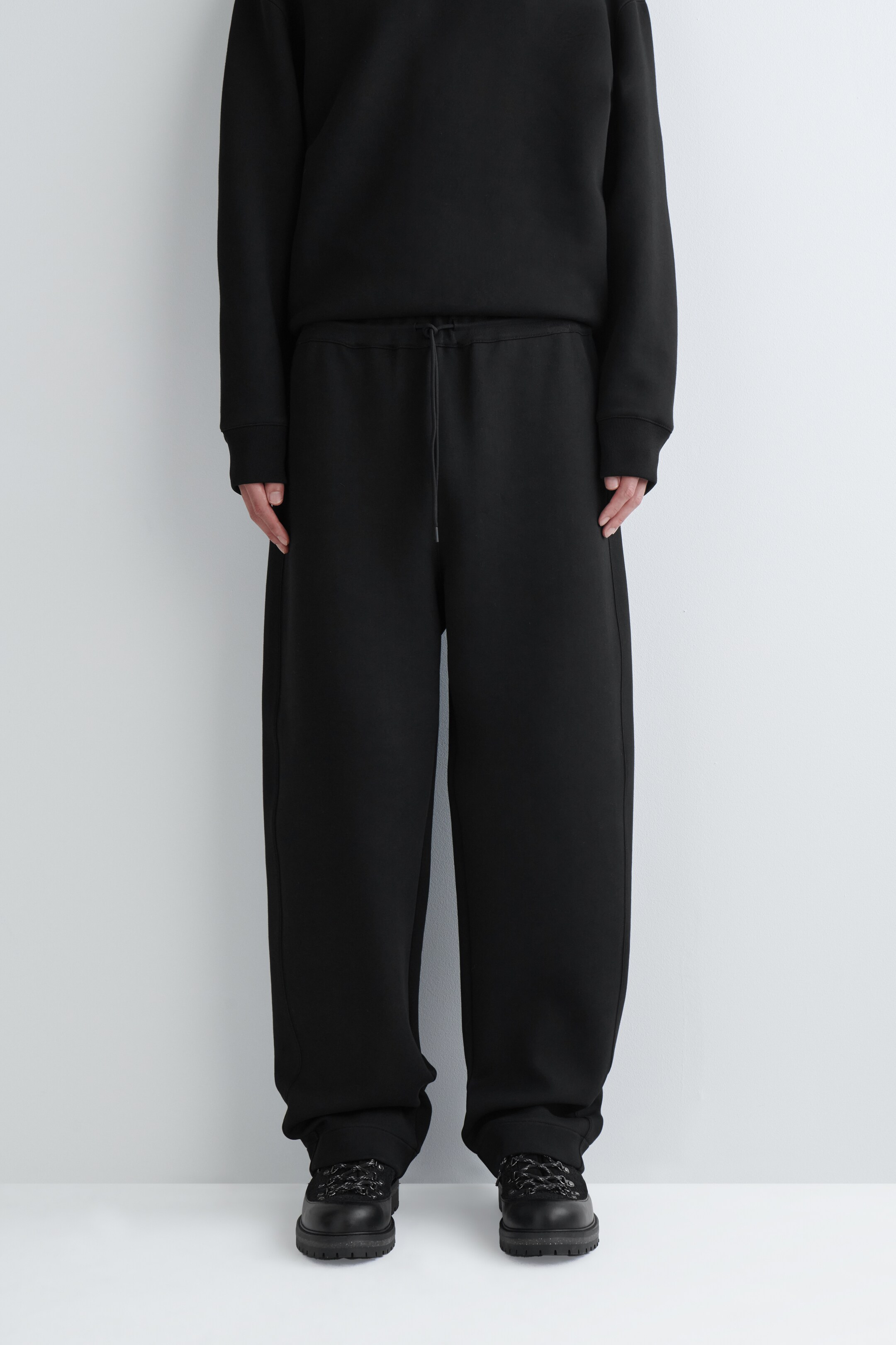 Agrandir l'image: Un pantalon noir ample est présenté sur une personne de face, avec une taille élastiquée et un cordon noir visible. Le pantalon présente une silhouette à jambes larges qui s'accumule légèrement sur des chaussures foncées.