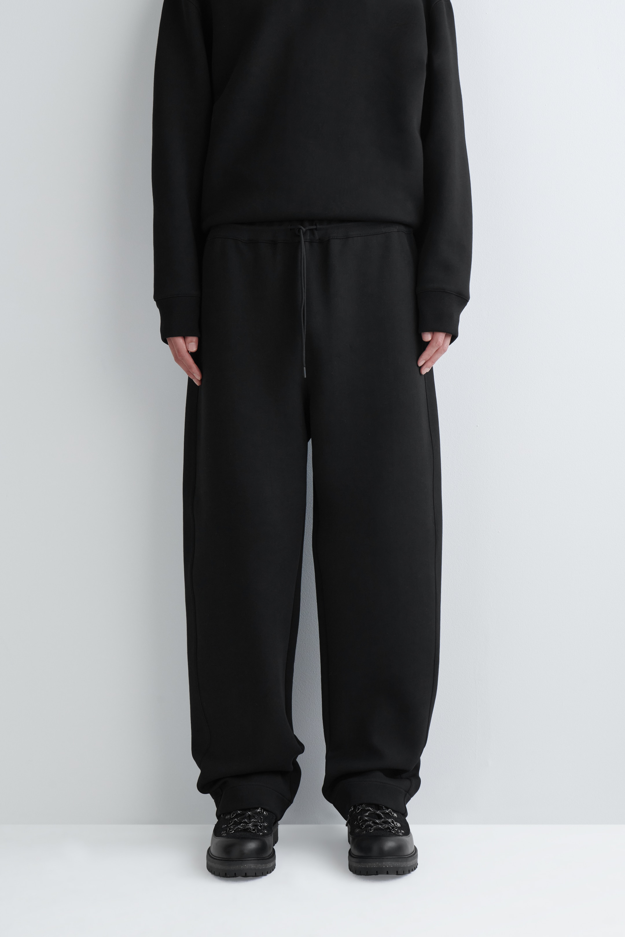 Vezi imaginea mai mare: O pereche de pantaloni negri, largi, este prezentată pe o persoană care stă cu fața înainte, având o talie elastică cu un șnur negru vizibil. Pantalonii prezintă o siluetă cu crac larg, care se adună ușor peste încălțămintea închisă la culoare.