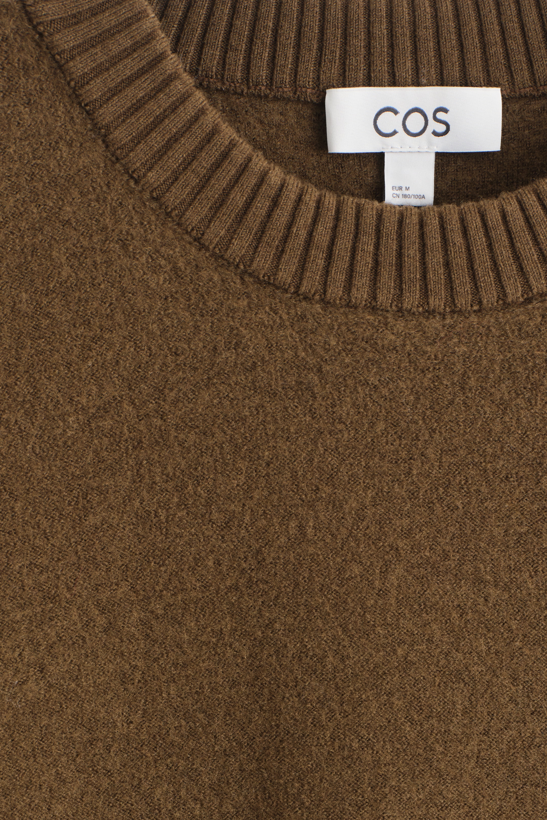 View larger image: A dark brown wool jumper is being presented from the front, showcasing a crew neck with a wide ribbed trim. A white brand label is visible at the inner neckline.