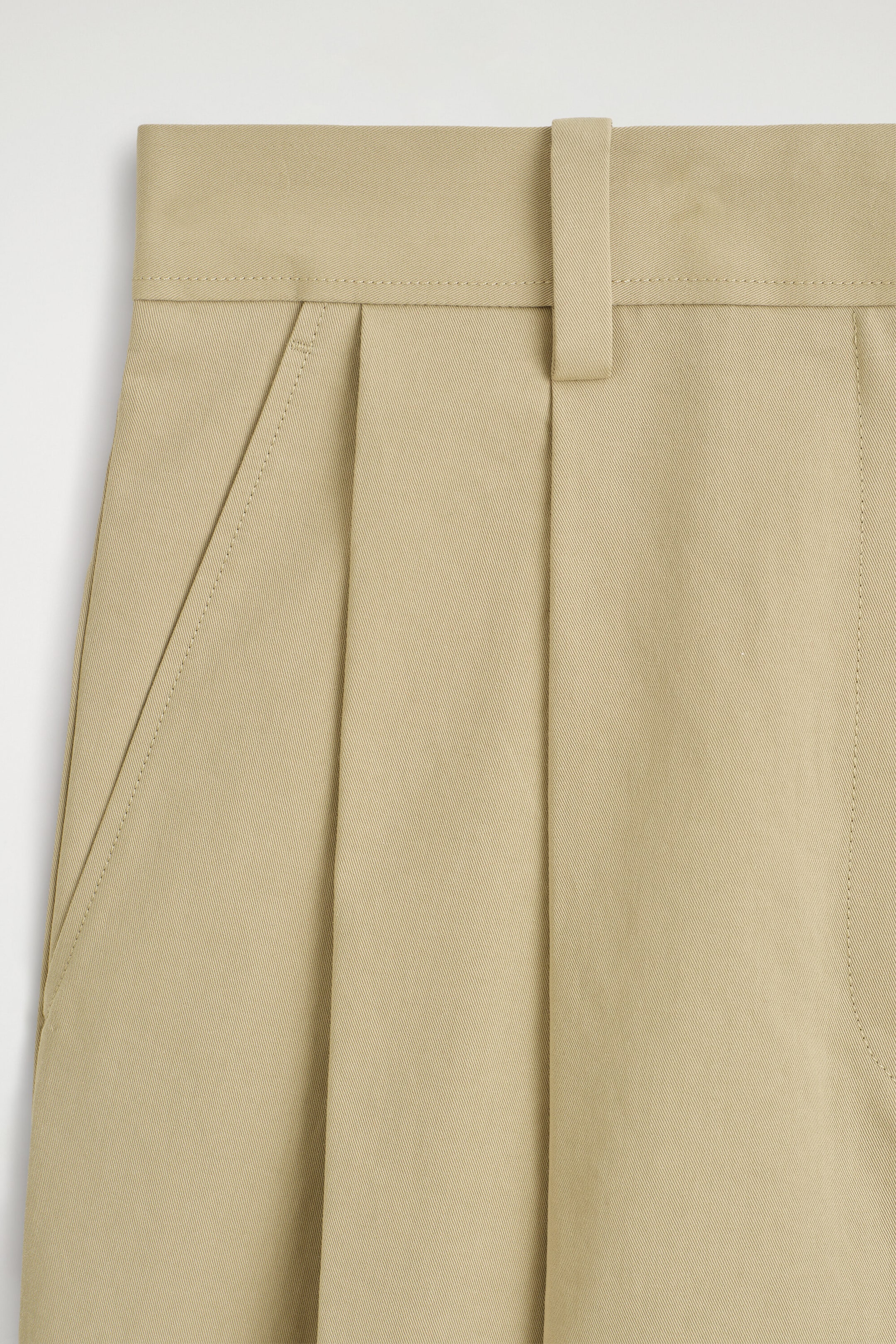 Showing a close-up front view of the cotton cropped barrel-leg pants in beige twill fabric. The design features a structured waistband with topstitching, a single wide belt loop, deep front pleats, and a slanted side pocket. Showing a close-up front view of the cotton cropped barrel-leg pants in beige twill fabric. The design features a structured waistband with topstitching, a single wide belt loop, deep front pleats, and a slanted side pocket.