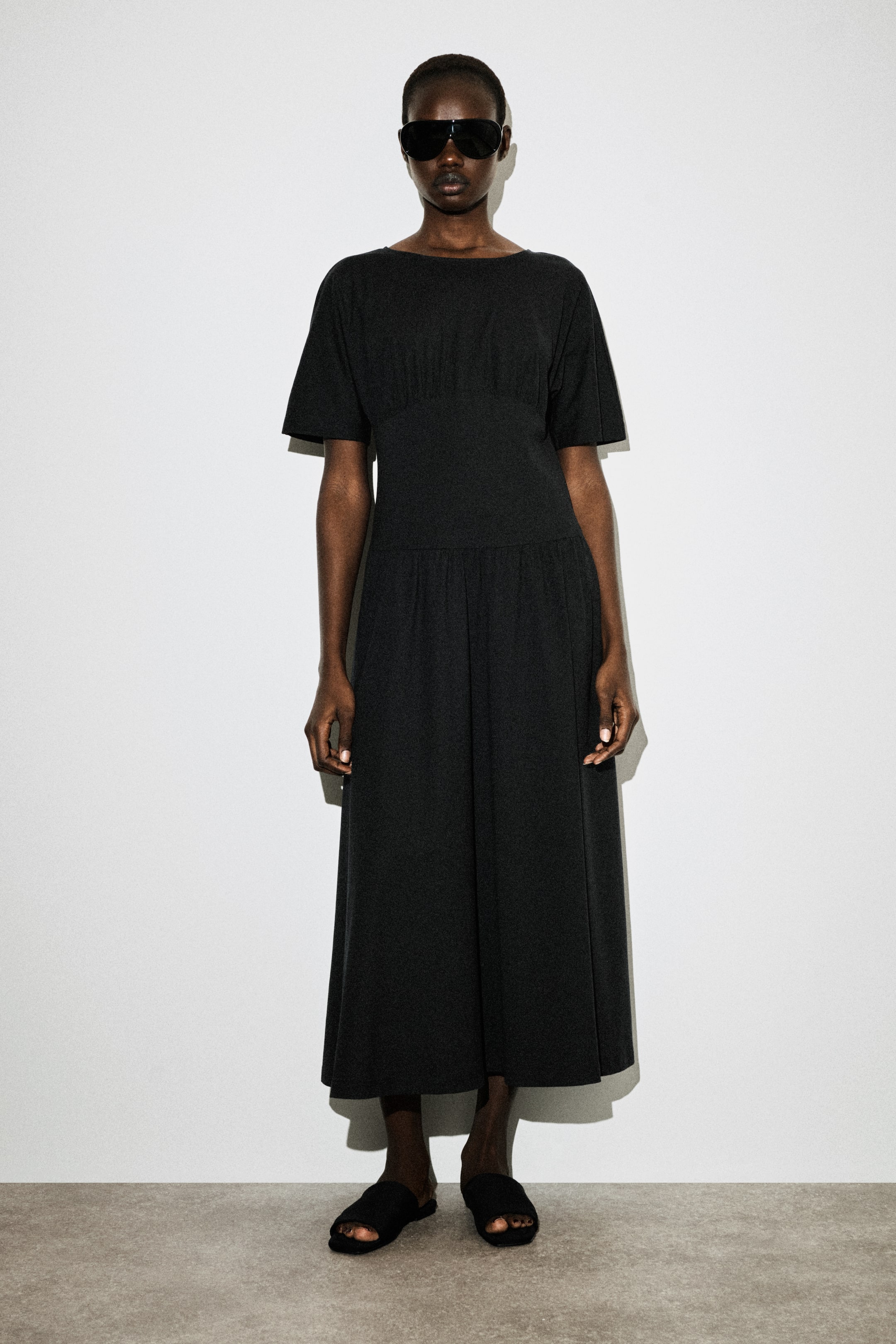 הצגת תמונה גדולה יותר: שמלה מידי שחורה עם מחשוף סירה, שרוולים קצרים וחלק עליון מכווץ מעל מותן צמודה. חצאית בגזרת A נופלת לאורך מידי.