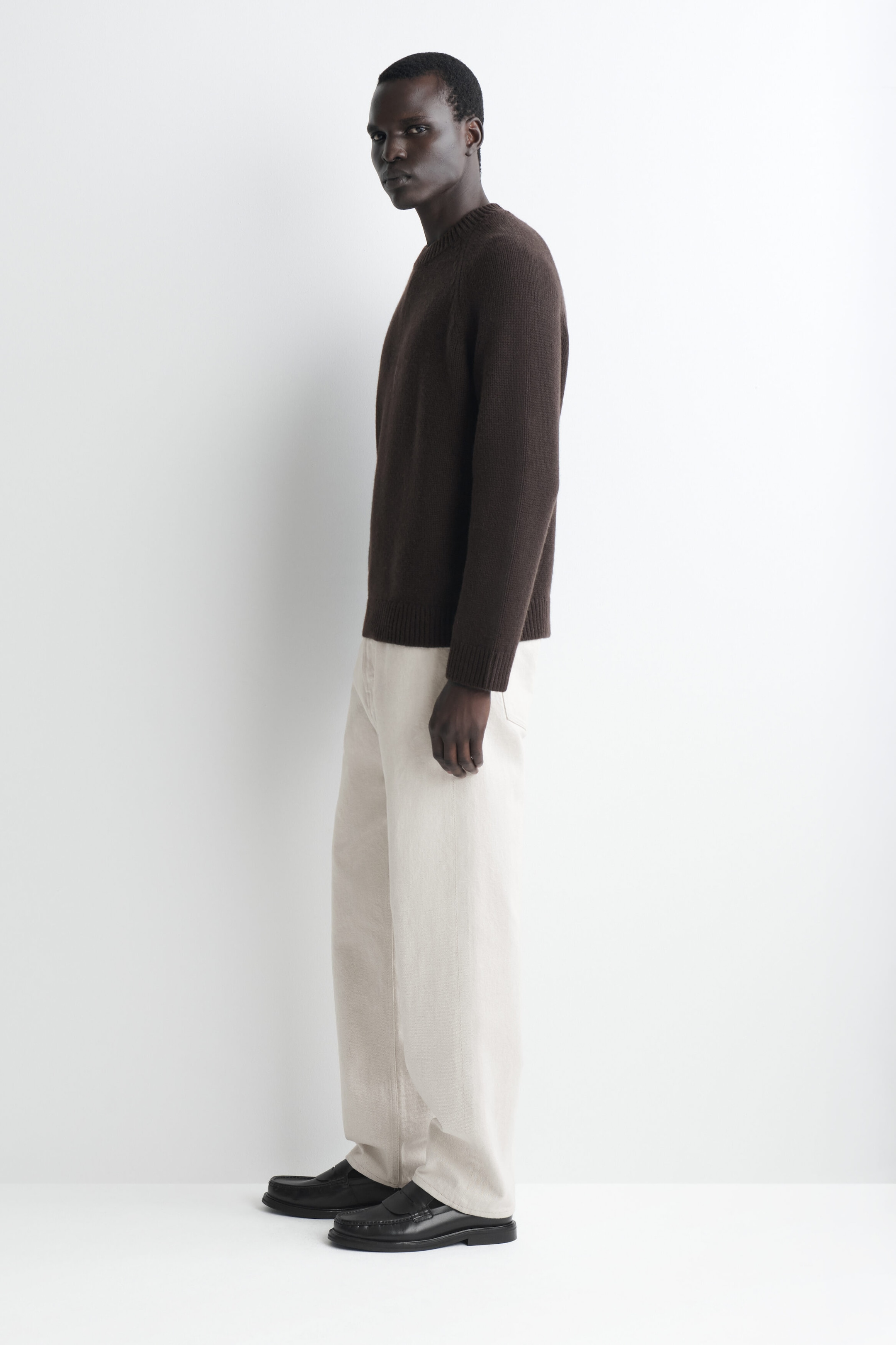 A dark brown chunky cashmere jumper is facing left in a side profile view, showcasing its relaxed fit, ribbed crew neck, ribbed cuffs, and ribbed hem. The jumper falls straight to the hips, where its hem rests over light-coloured trousers. A dark brown chunky cashmere jumper is facing left in a side profile view, showcasing its relaxed fit, ribbed crew neck, ribbed cuffs, and ribbed hem. The jumper falls straight to the hips, where its hem rests over light-coloured trousers.