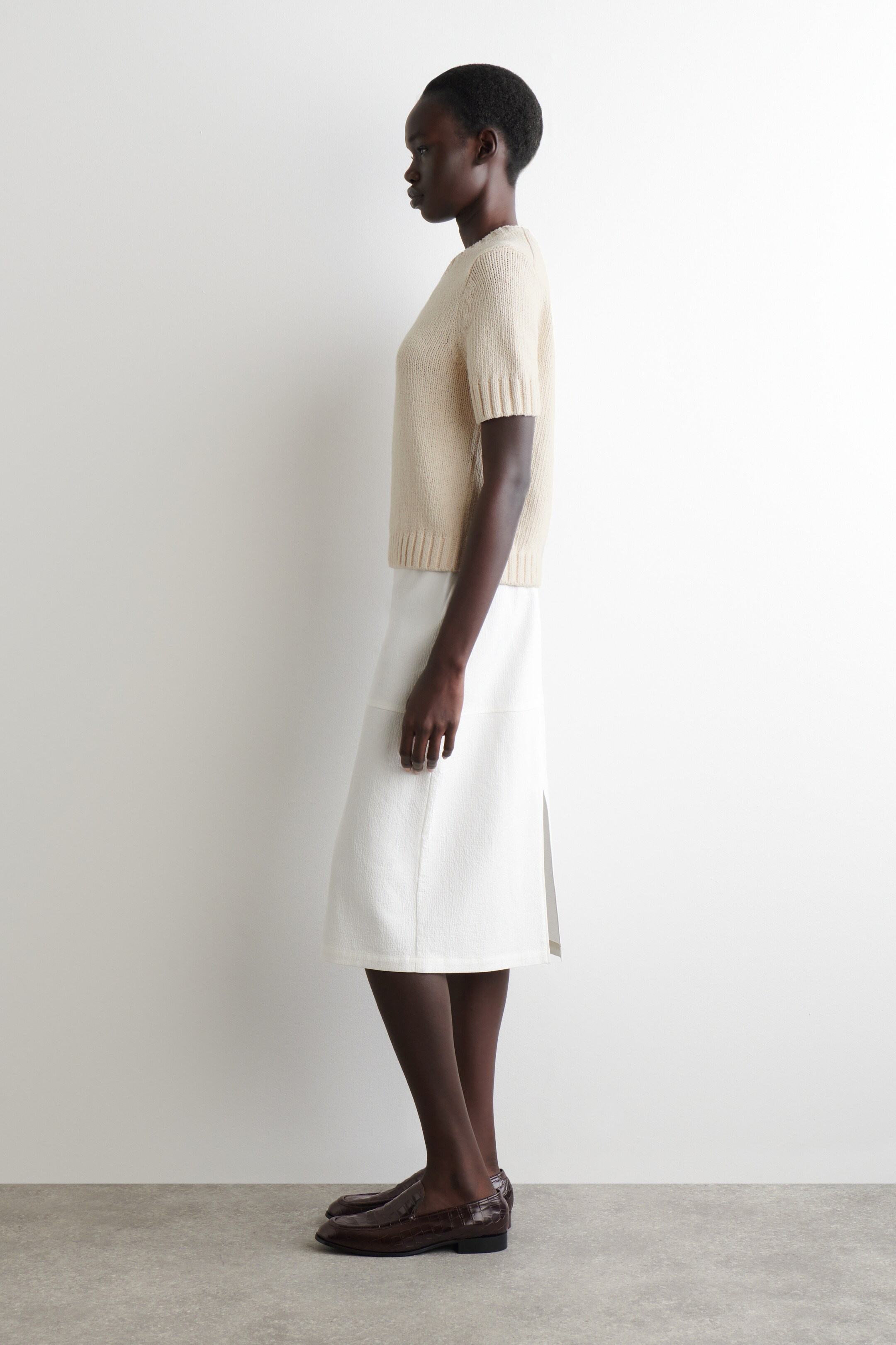 View larger image: A light beige knit short-sleeved top with ribbed cuffs and hem is being worn, showing a relaxed fit from the side. It is being paired with a white textured midi skirt featuring a side seam and slit, and dark brown embossed loafers are completing the ensemble.
