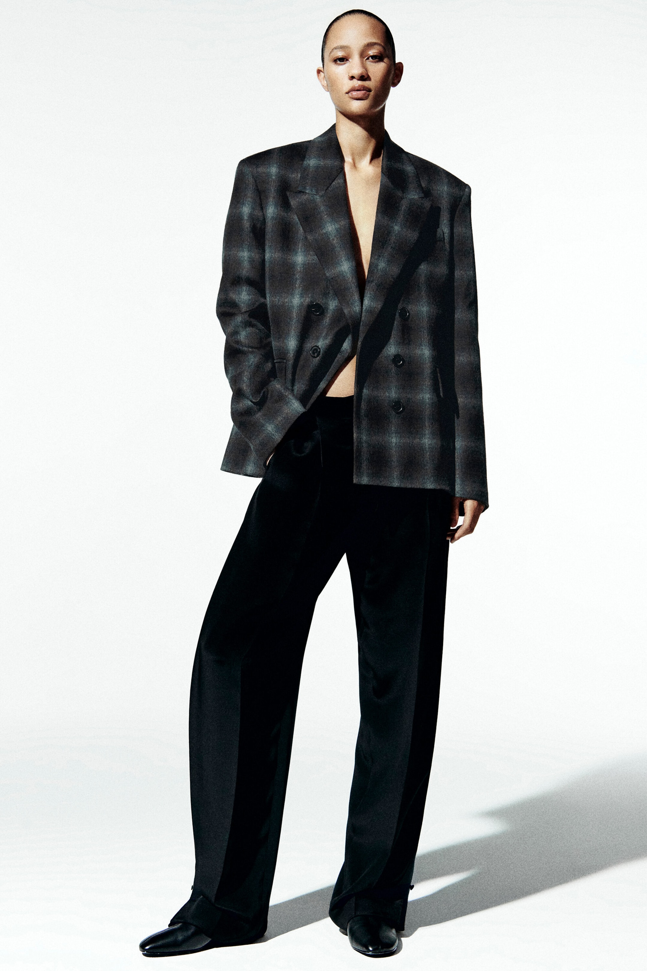 Un blazer à double boutonnage avec un motif à carreaux diffus gris foncé et noir est présenté de face, avec une coupe décontractée, des revers en pointe et une fermeture à boutons. Le blazer est porté ouvert sur un pantalon noir à jambes larges.