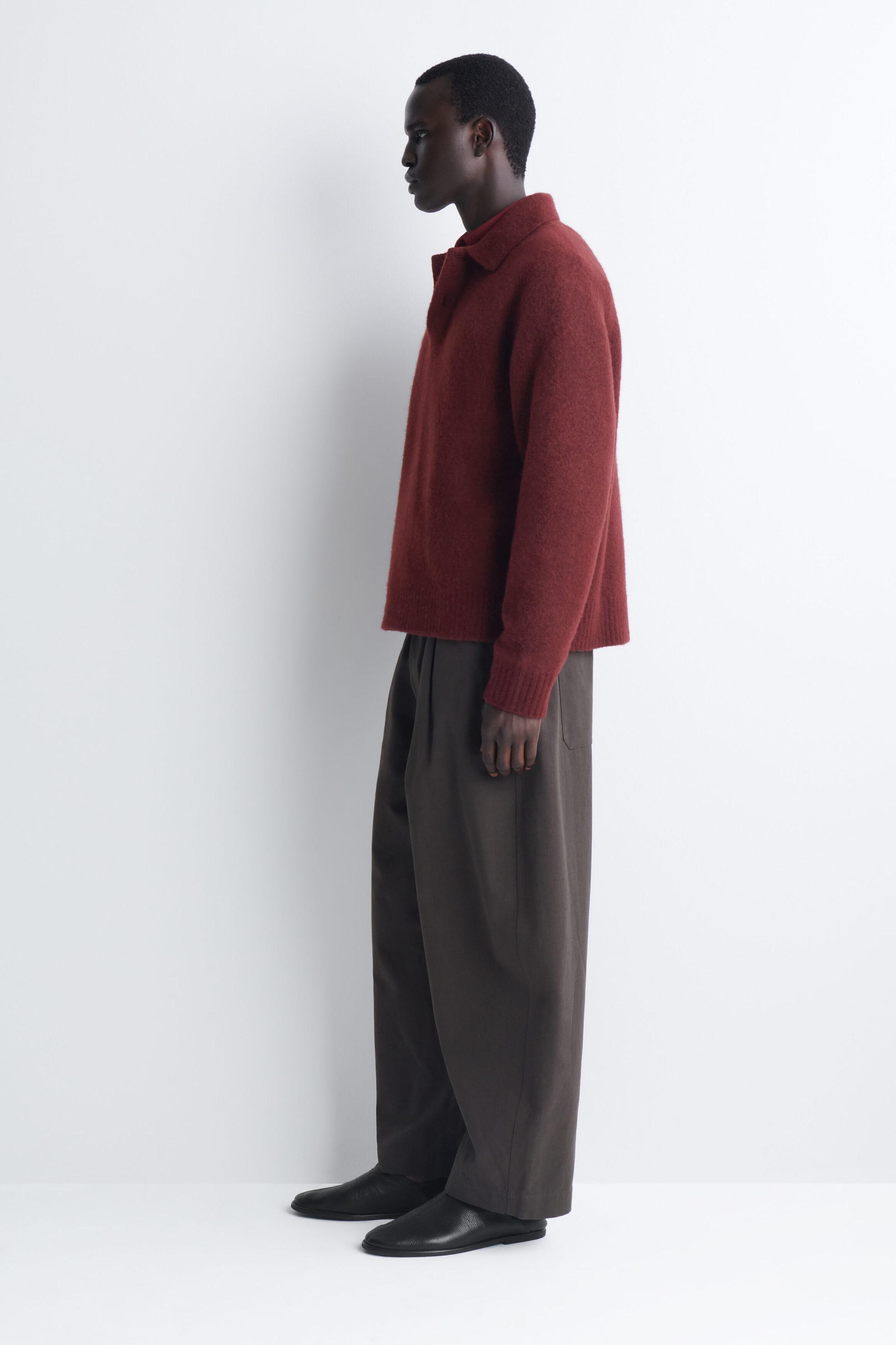 The brushed-wool polo shirt in a deep rust-red color is shown from a left-facing side profile, highlighting its relaxed fit and soft, fuzzy texture. The shirt features a polo collar and ribbed cuffs, and drapes loosely over dark brown trousers. The brushed-wool polo shirt in a deep rust-red color is shown from a left-facing side profile, highlighting its relaxed fit and soft, fuzzy texture. The shirt features a polo collar and ribbed cuffs, and drapes loosely over dark brown trousers.