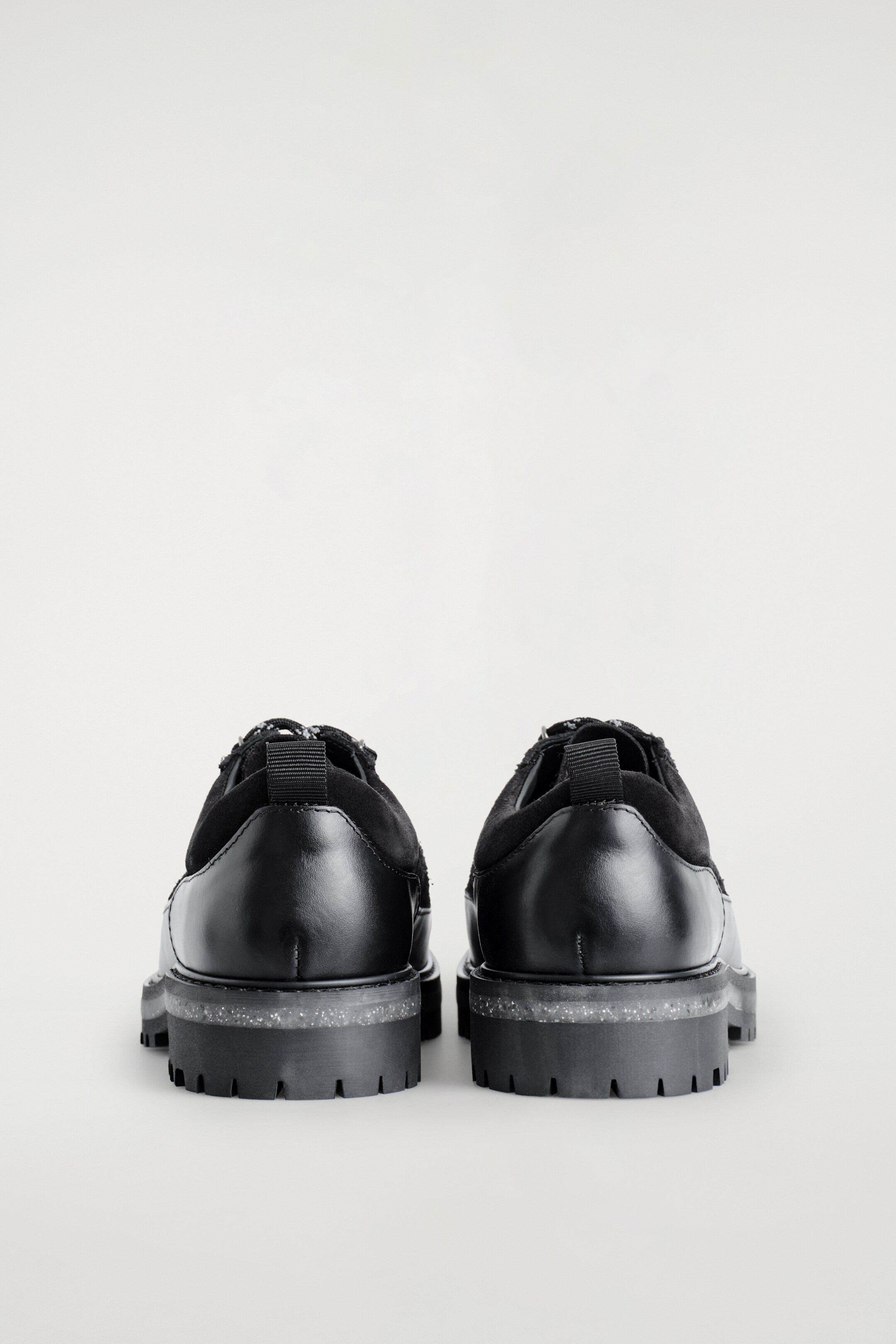 Ver imagen más grande: Un par de zapatos negros se están viendo desde atrás. Presentan cuero liso en el talón y ante cepillado alrededor del tobillo, con una lengüeta de tirón negra visible. Los zapatos descansan sobre suelas gruesas y en capas que incluyen una capa intermedia translúcida y moteada y una suela exterior negra con tacos.
