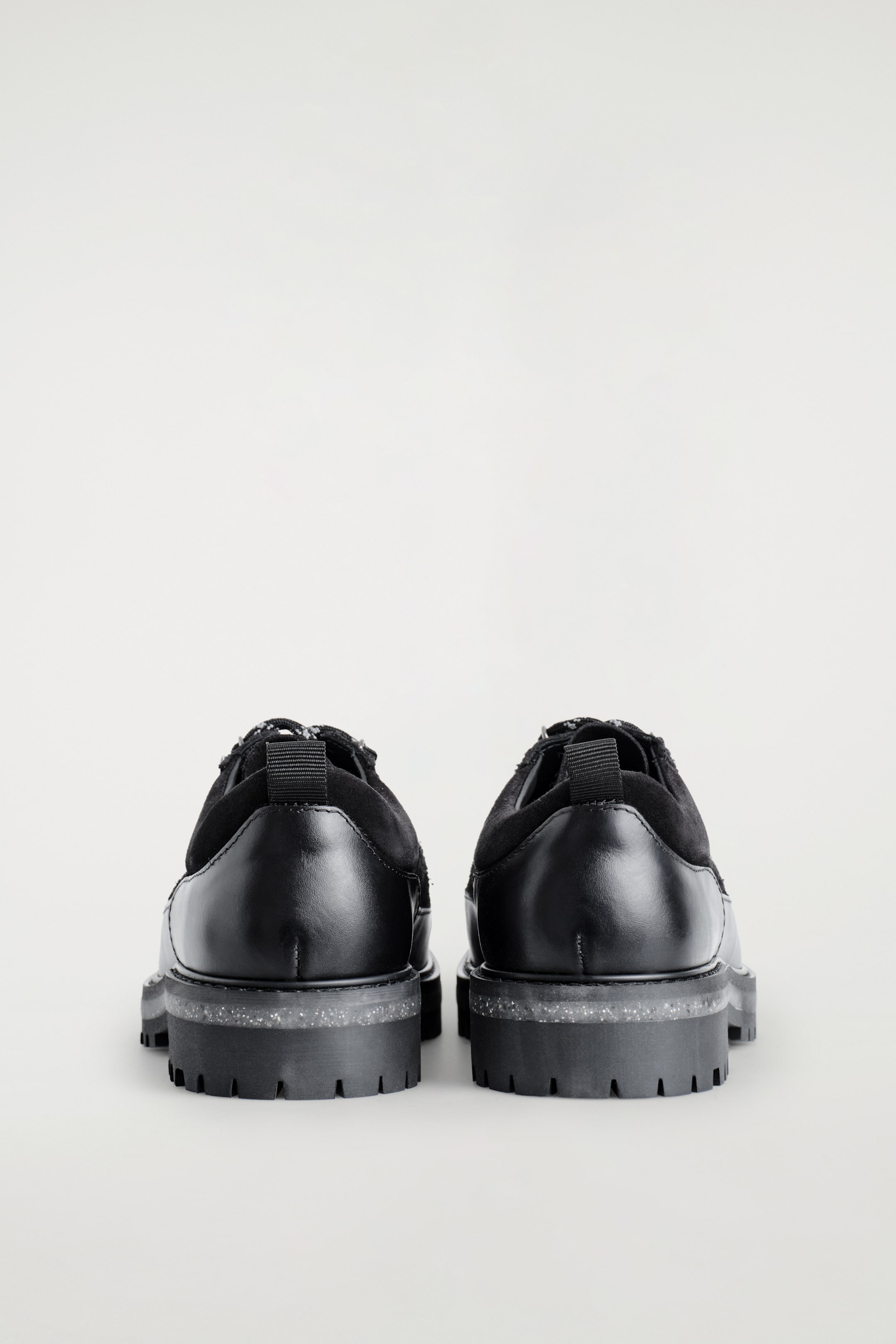 Ver imagem maior: Um par de sapatos pretos está a ser visto de trás. Eles apresentam couro liso no calcanhar e camurça escovada em torno do tornozelo, com uma aba de puxar preta visível. Os sapatos estão apoiados em solas grossas e em camadas que incluem uma camada intermédia translúcida e salpicada e uma sola exterior preta com ranhuras.