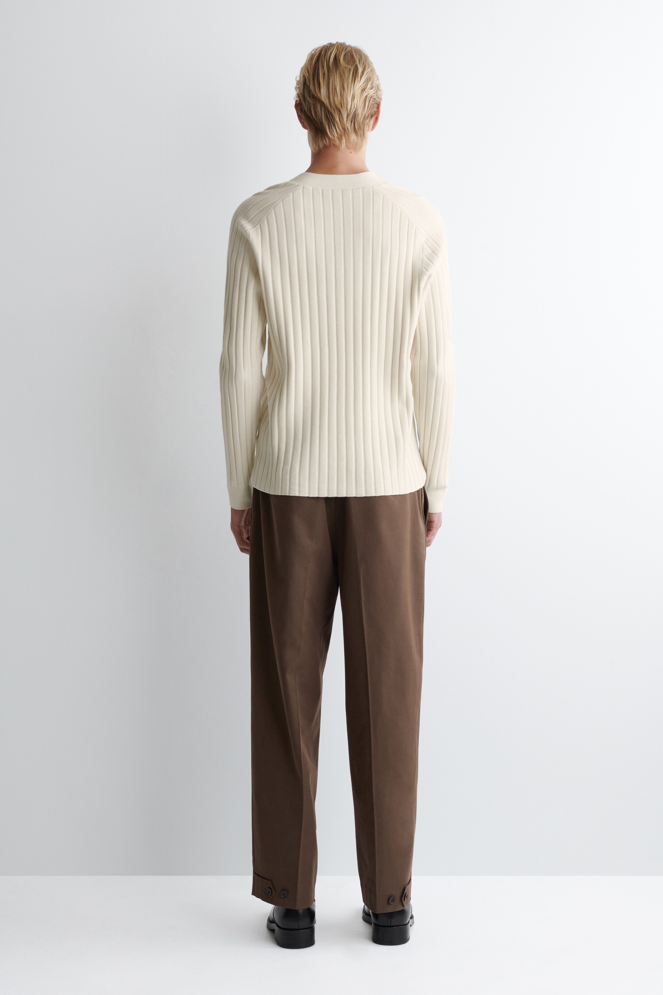Agrandir l'image: Un pull en maille côtelée crème est présenté de dos, avec des manches raglan et un col rond. Il est assorti à un pantalon marron à coupe décontractée, avec des détails de pattes boutonnées près de la cheville et des chaussures de ville noires.