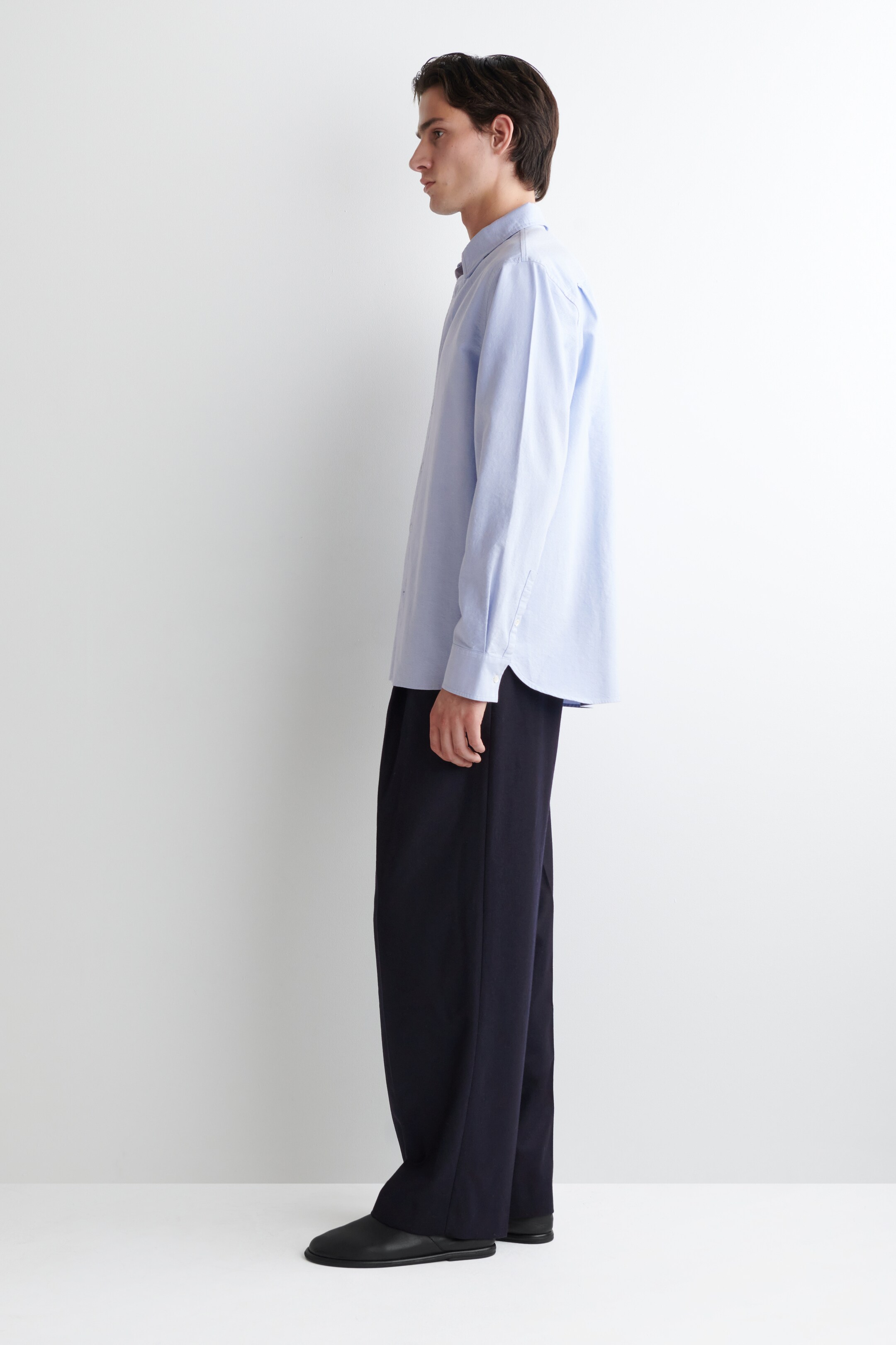 Ver imagen más grande: Una camisa azul claro de botones, con un ajuste relajado, mangas largas y puños abotonados, se lleva con pantalones anchos de color azul oscuro que presentan una silueta larga y fluida. Este conjunto, completado con zapatos negros sin cordones, se muestra de perfil lateral, mirando hacia la izquierda.