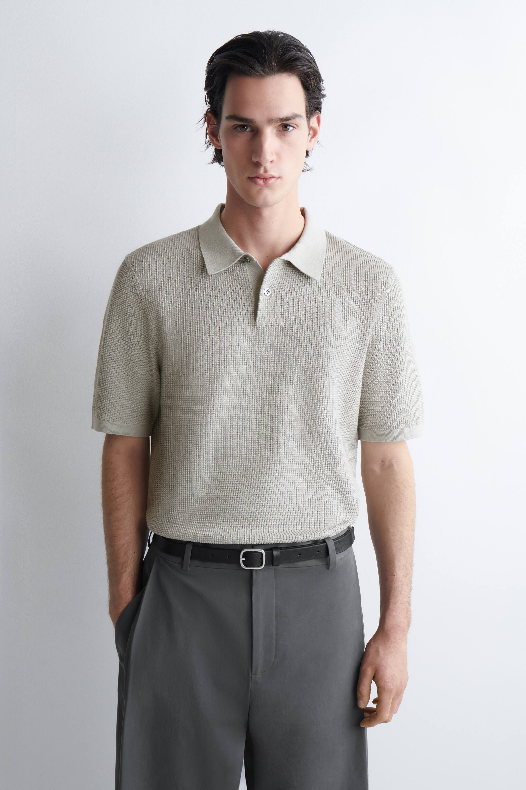 Näytä suurempi kuva: Vaaleanbeige, teksturoitu neulepaitapusero esitellään, jossa on klassinen kaulus, kaksinappinen nappilista ja lyhyet hihat. Se on puettu tummien housujen sisään.