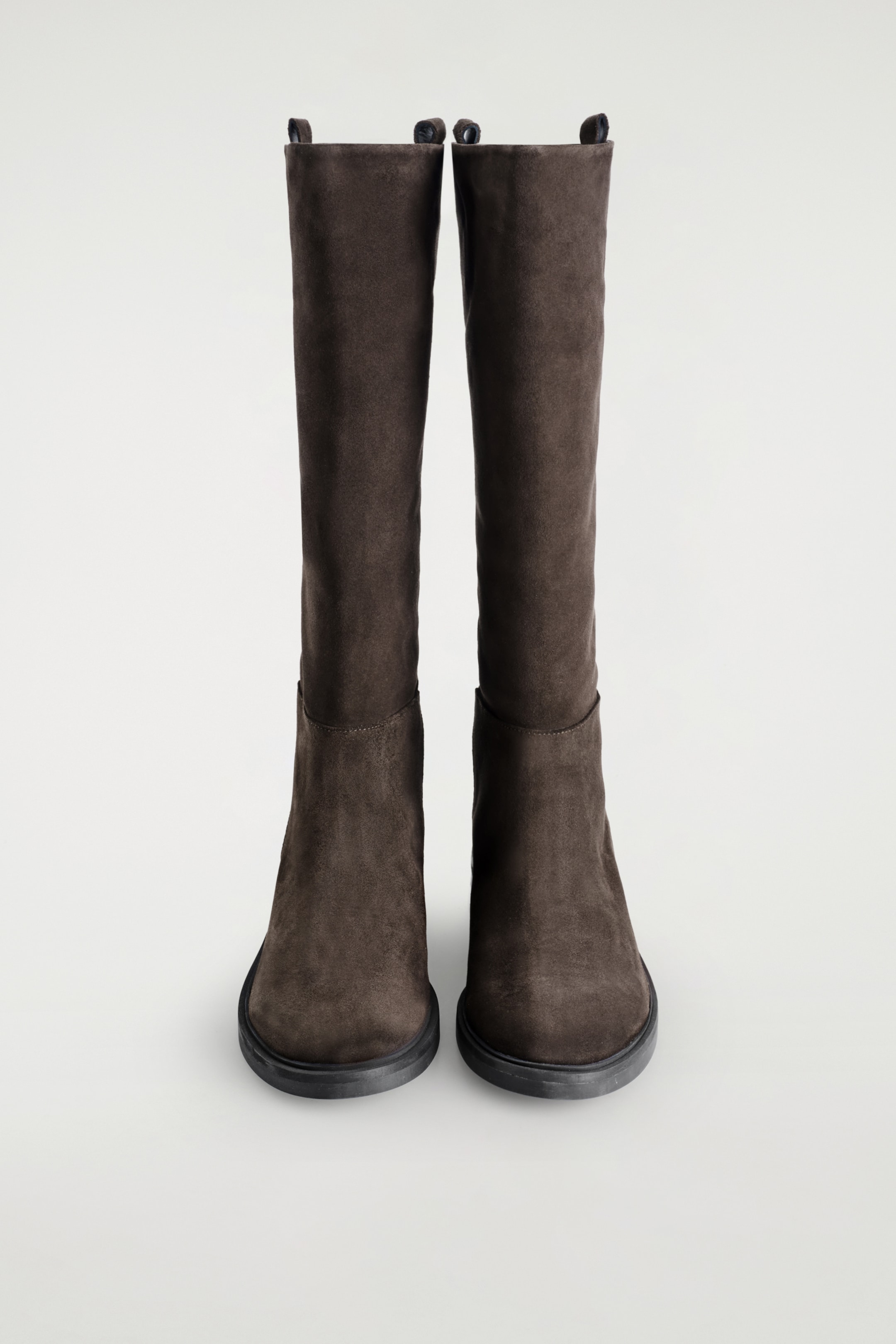 Größeres Bild ansehen: Ein Paar dunkelbraune Wildlederstiefel steht frontseitig, mit glatten Schäften, abgerundeten Zehen und flachen dunklen Sohlen. Dunkle Zuglaschen sind an der oberen Öffnung jedes Stiefels sichtbar.