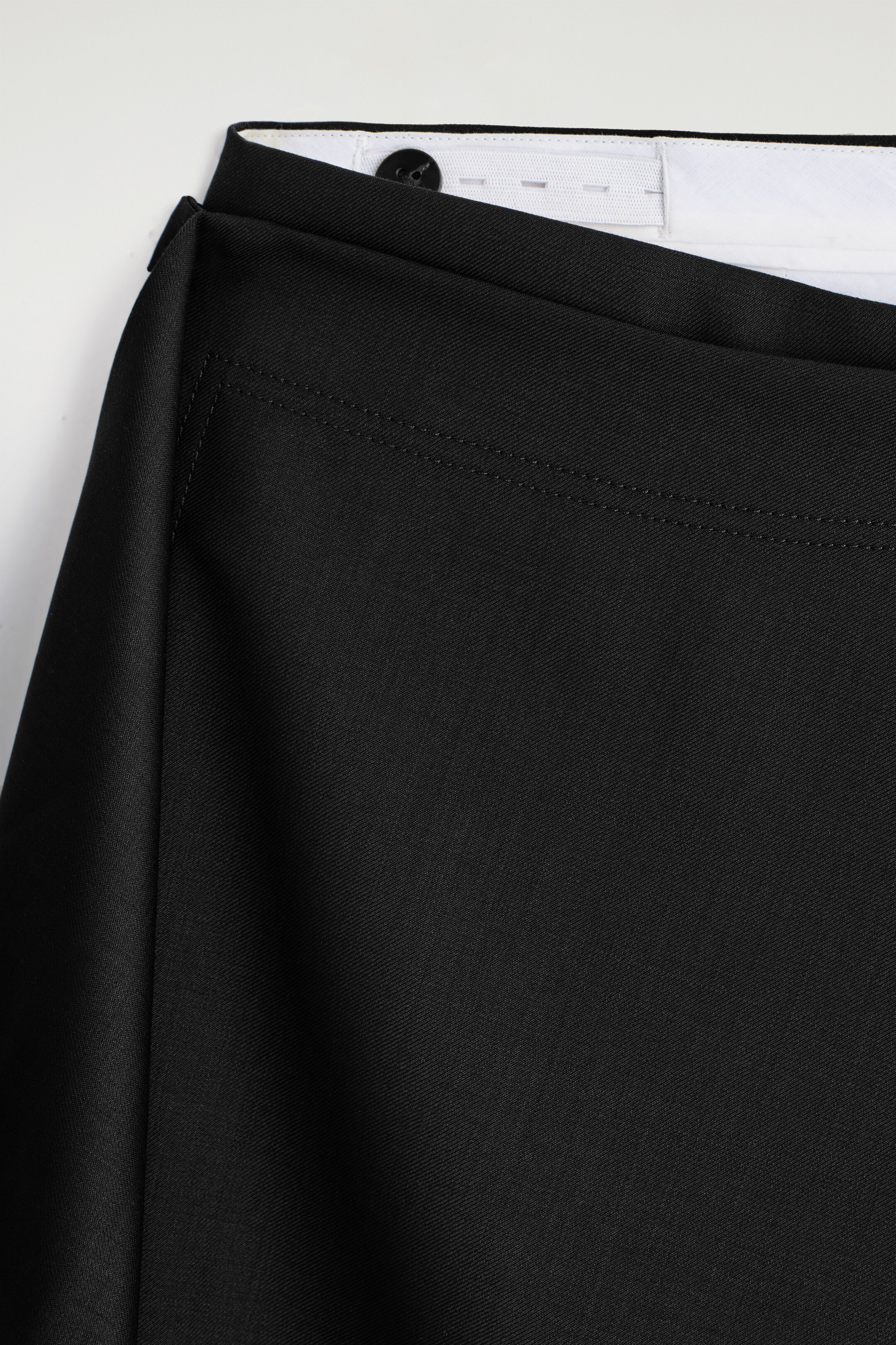 Agrandir l'image: Un gros plan de pantalon noir montre la partie supérieure avant, avec un panneau drapé à la taille et une double surpiqûre. Une ceinture intérieure blanche avec un bouton noir et une fermeture à crochet est visible.