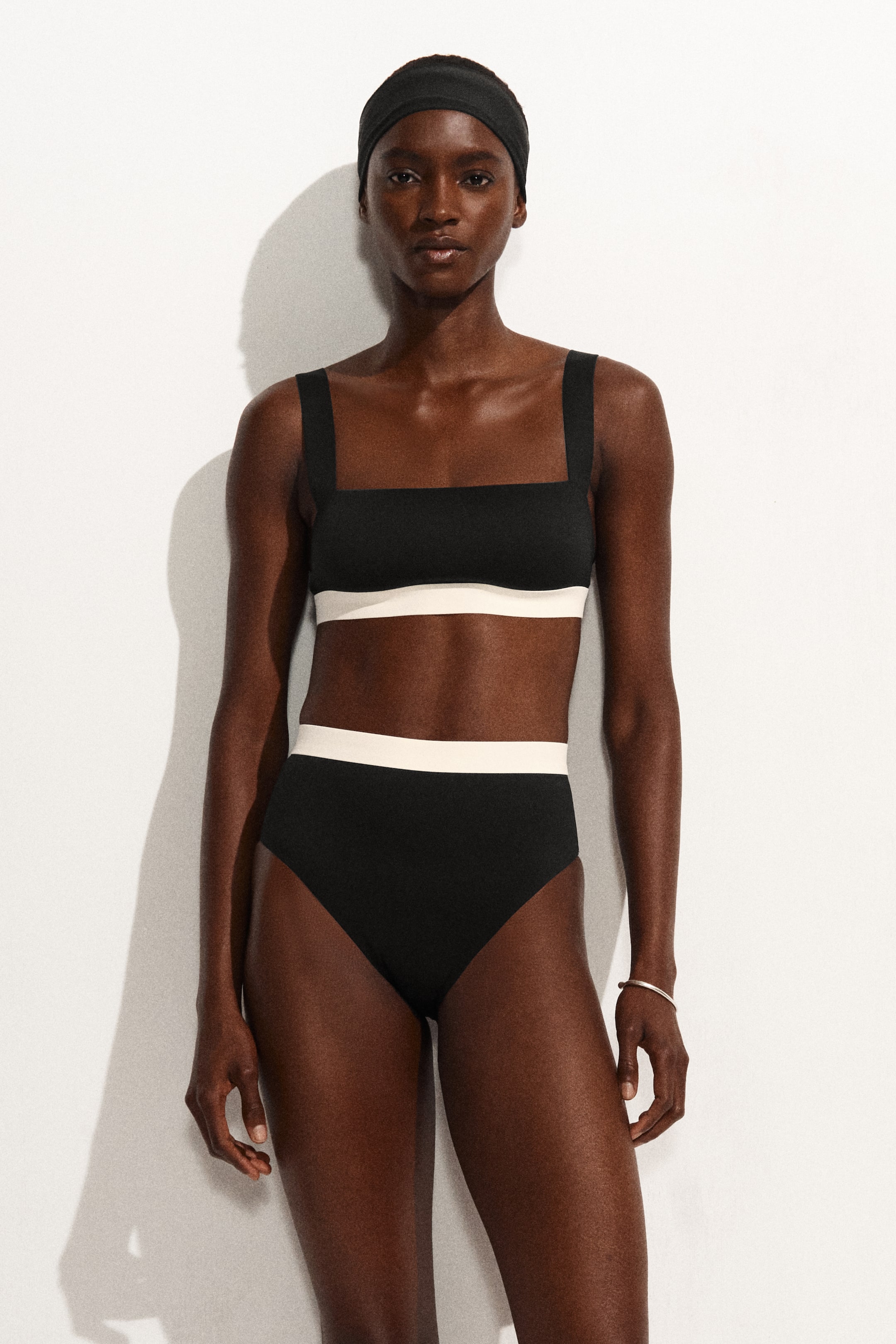 Grotere afbeelding bekijken: Een tweedelig badpak met een zwarte vierkante hals bikini top met brede bandjes en een witte horizontale band, gecombineerd met bijpassende zwarte hoog getailleerde bikinibroekjes die ook een witte horizontale tailleband hebben. Het badpak is naar voren gericht, waardoor het tweekleurige ontwerp wordt benadrukt.