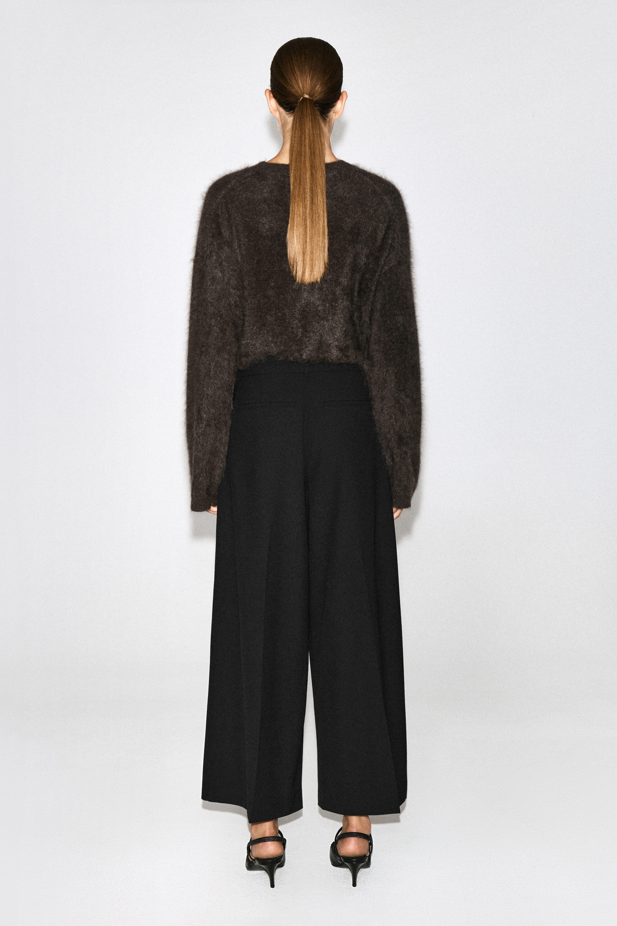 Pleated wool-blend wide-leg trousers in black are worn by the model, viewed from the back to show the high-waisted fit and relaxed silhouette. The trousers feature a sharp pressed crease running down the back of each leg and are styled with a tucked-in, dark brown textured jumper. Pleated wool-blend wide-leg trousers in black are worn by the model, viewed from the back to show the high-waisted fit and relaxed silhouette. The trousers feature a sharp pressed crease running down the back of each leg and are styled with a tucked-in, dark brown textured jumper.