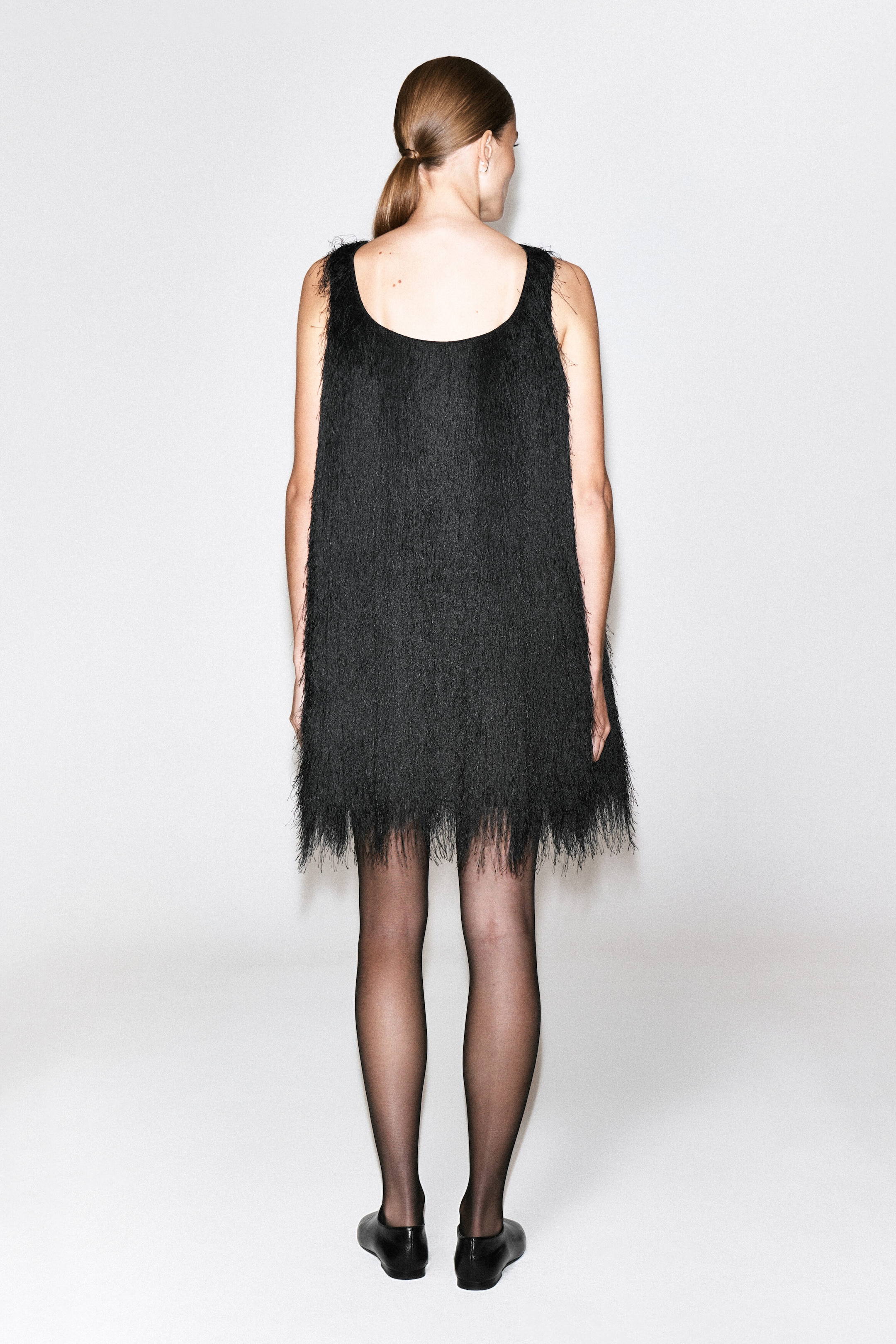 Ein schwarzes Mini-Kleid mit Fransen, ärmellosem Design, tiefem Rückenausschnitt und dichtem Fransenbesatz über das gesamte Kleid wird vom Model getragen. Das Kleid wird mit schwarzen, transparenten Strumpfhosen und flachen schwarzen Schuhen kombiniert. Ein schwarzes Mini-Kleid mit Fransen, ärmellosem Design, tiefem Rückenausschnitt und dichtem Fransenbesatz über das gesamte Kleid wird vom Model getragen. Das Kleid wird mit schwarzen, transparenten Strumpfhosen und flachen schwarzen Schuhen kombiniert.