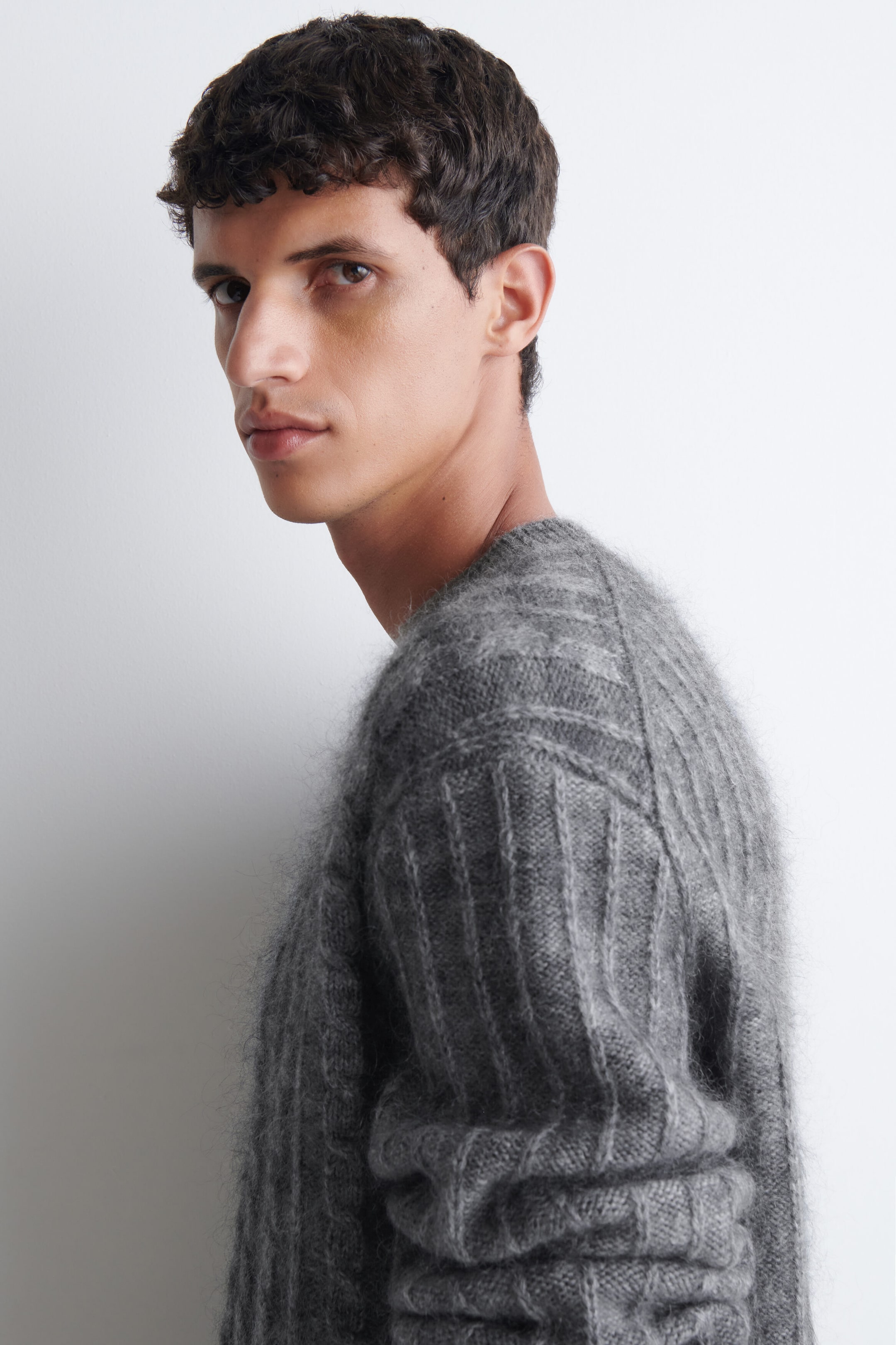 A grey, fuzzy rib-knit sweater is being worn by the model, who is turned in profile. The sweater features a textured, soft surface with vertical ribbing along the sleeve and body. A grey, fuzzy rib-knit sweater is being worn by the model, who is turned in profile. The sweater features a textured, soft surface with vertical ribbing along the sleeve and body.