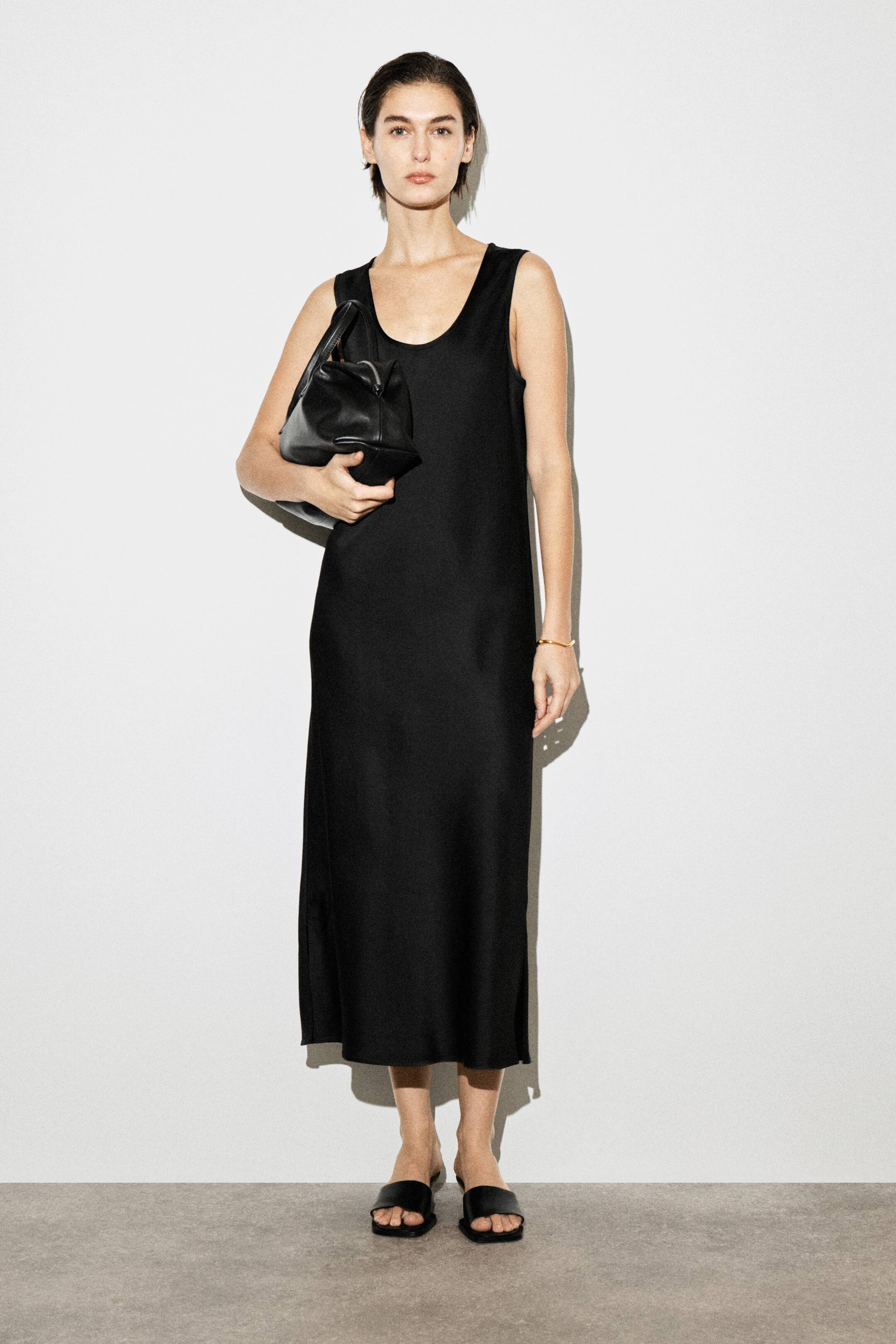 Größeres Bild anzeigen: Ein schwarzes ärmelloses Midikleid mit Rundhalsausschnitt wird von dem Model getragen, das auch eine schwarze Handtasche trägt und schwarze flache Sandalen anhat.