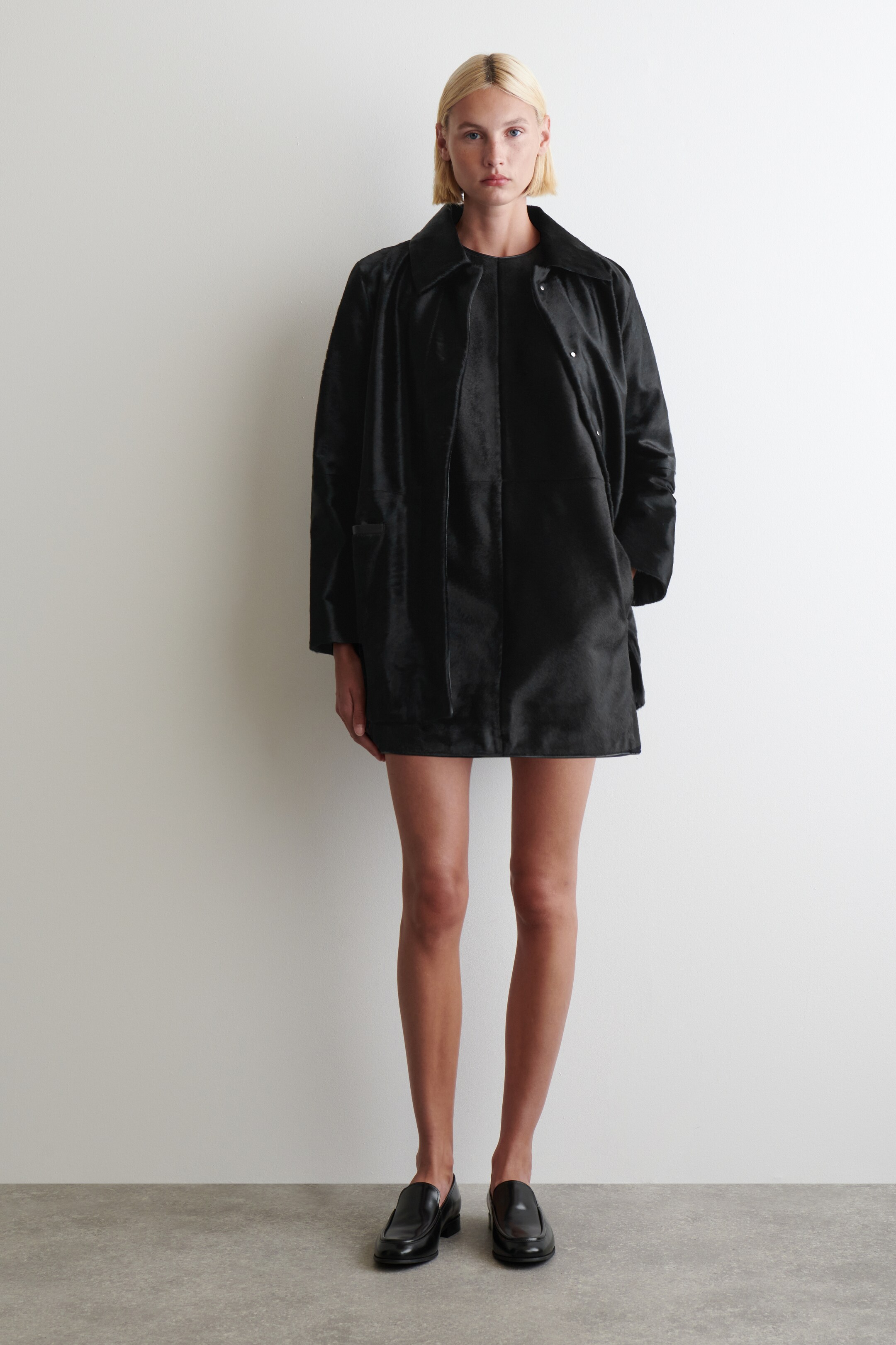 Agrandir l'image: Un manteau noir à texture courte est présenté, avec un large col plat, une patte de boutonnage cachée avec des pressions apparentes de ton argenté en haut, et deux grandes poches avant. Le manteau ample, de longueur mi-cuisse, est porté ouvert sur un vêtement noir.