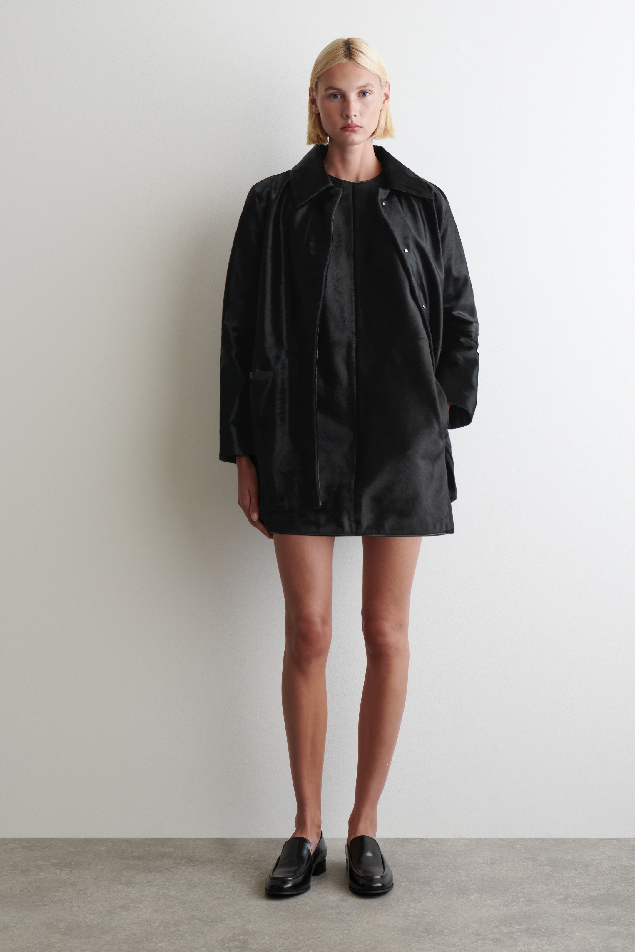 Agrandir l'image: Un manteau noir à texture courte est présenté, avec un large col plat, une patte de boutonnage cachée avec des pressions apparentes de ton argenté en haut, et deux grandes poches avant. Le manteau ample, de longueur mi-cuisse, est porté ouvert sur un vêtement noir.