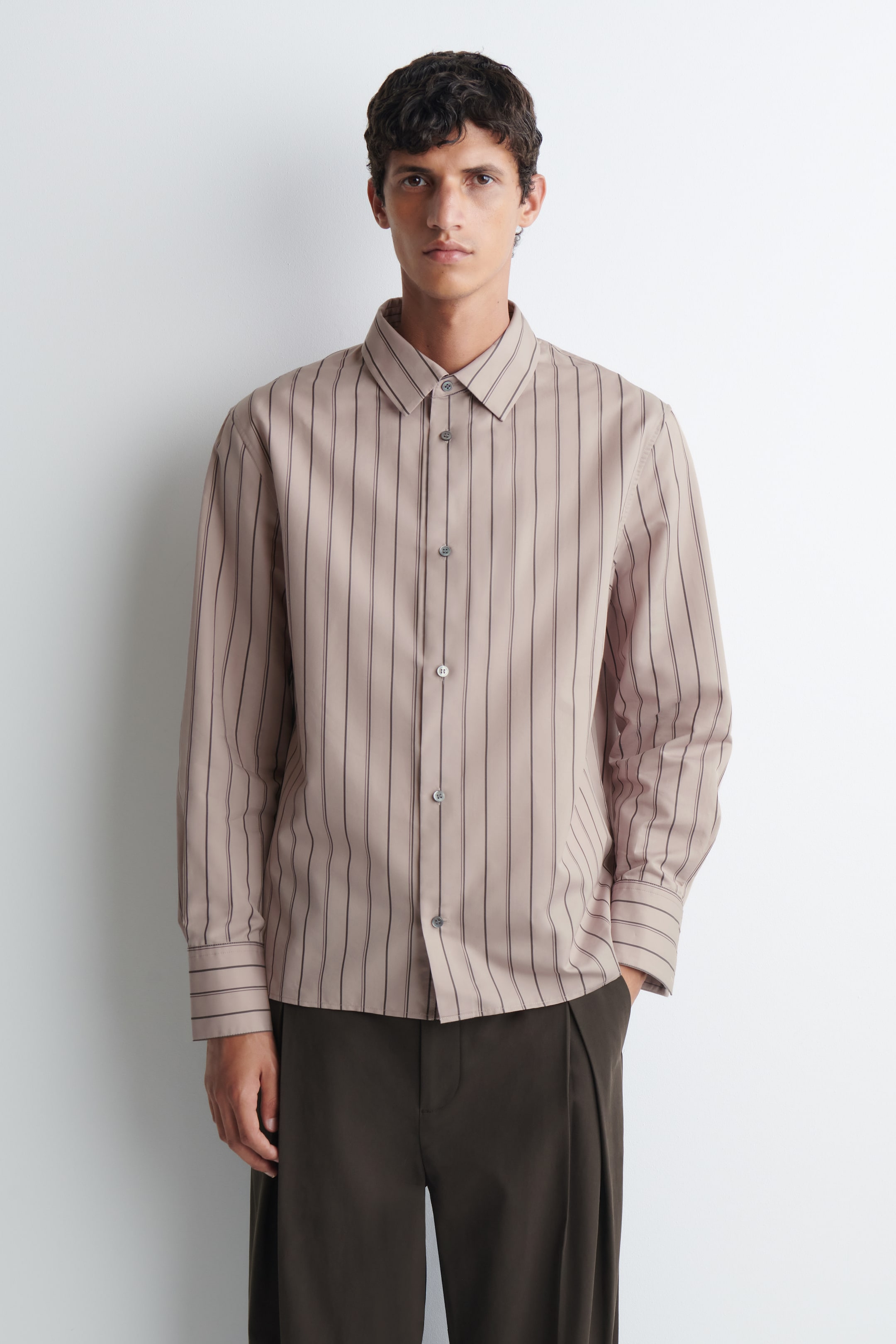 Agrandir l'image: Une chemise structurée en coton et soie à rayures, dans une teinte taupe clair avec de fines rayures verticales foncées, est portée par le mannequin. La chemise présente un col pointu, une fermeture boutonnée sur le devant et de larges poignets, et est portée rentrée à l’extérieur sur un pantalon plissé marron foncé.