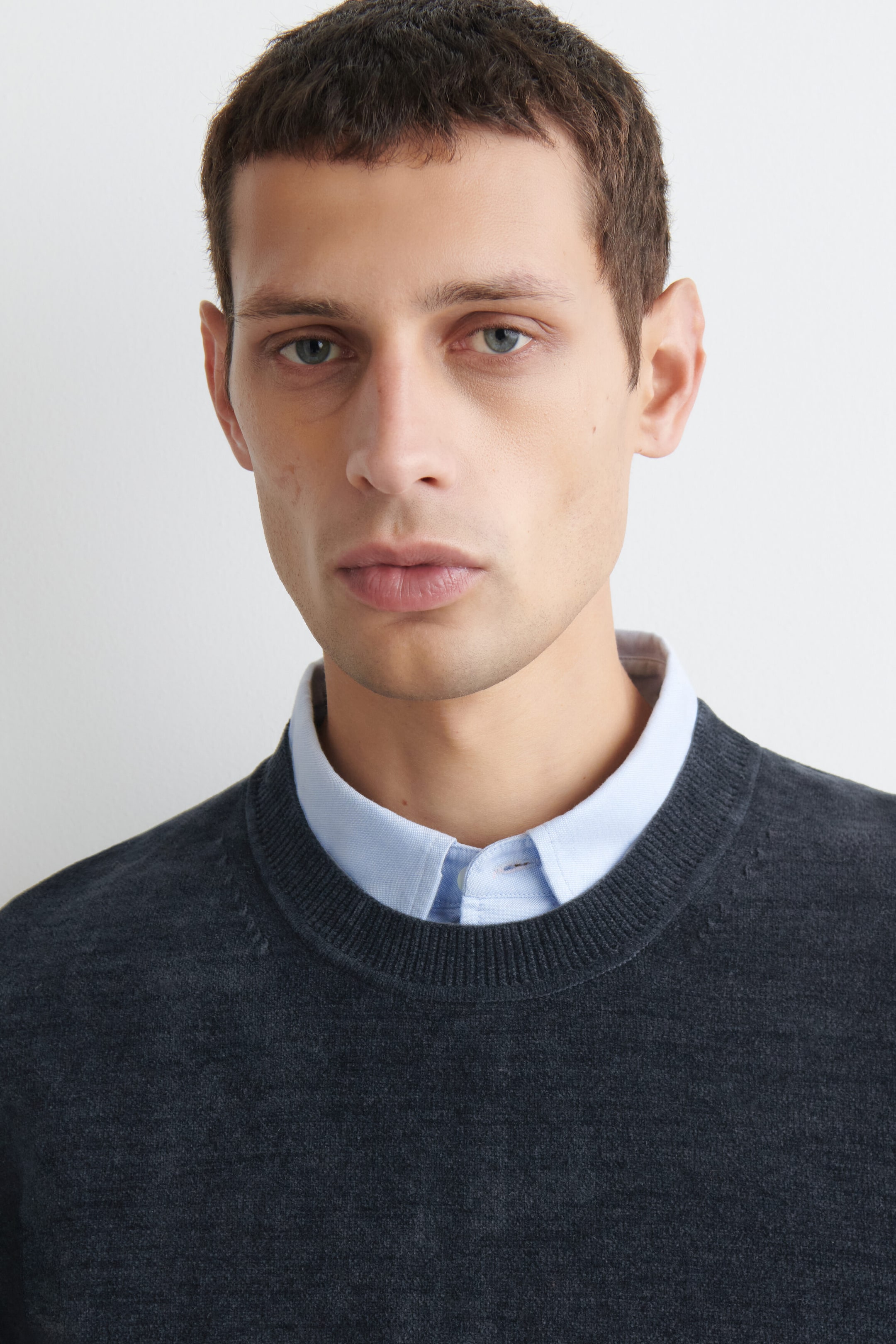 A dark gray cotton-chenille crewneck sweater is shown in a close-up front view, featuring a finely ribbed neckline and a soft, textured knit finish. The sweater is layered over a light blue button-down collared shirt to highlight its classic crewneck neckline. A dark gray cotton-chenille crewneck sweater is shown in a close-up front view, featuring a finely ribbed neckline and a soft, textured knit finish. The sweater is layered over a light blue button-down collared shirt to highlight its classic crewneck neckline.