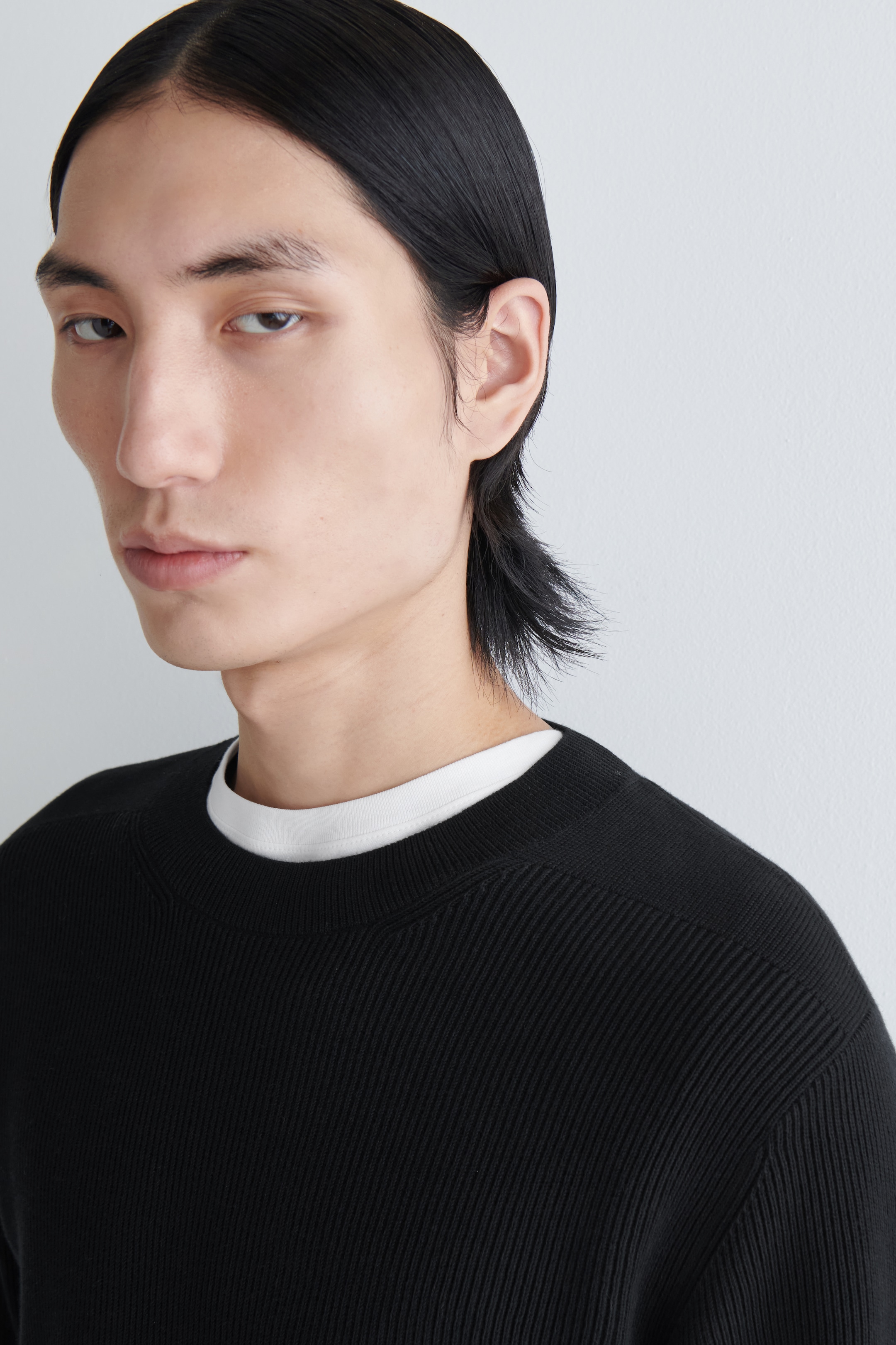 Agrandir l'image: Un pull noir en tricot côtelé est porté, présenté sous un angle légèrement avant-droit, avec un col rond, un motif de côtes verticales distinct et des détails de tricot subtils aux épaules. Un t-shirt blanc à col rond est superposé en dessous, dépassant légèrement au niveau de l'encolure.