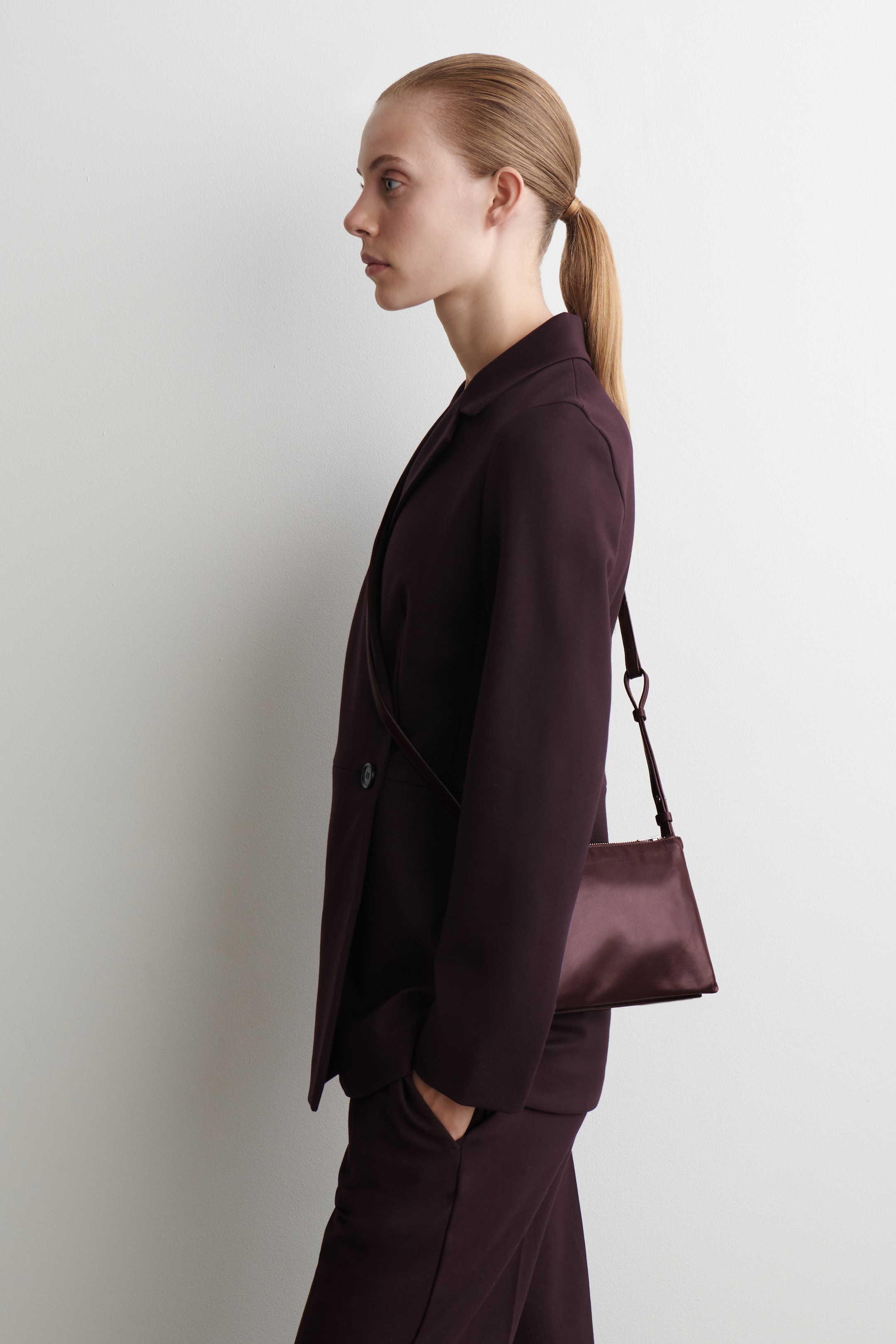 Agrandir l'image: Un tailleur-pantalon violet foncé est présenté de profil. Le costume comprend un blazer avec des revers crantés et un pantalon assorti. Un petit sac à bandoulière violet foncé et lustré avec une fine sangle est porté, pendu à la hanche.