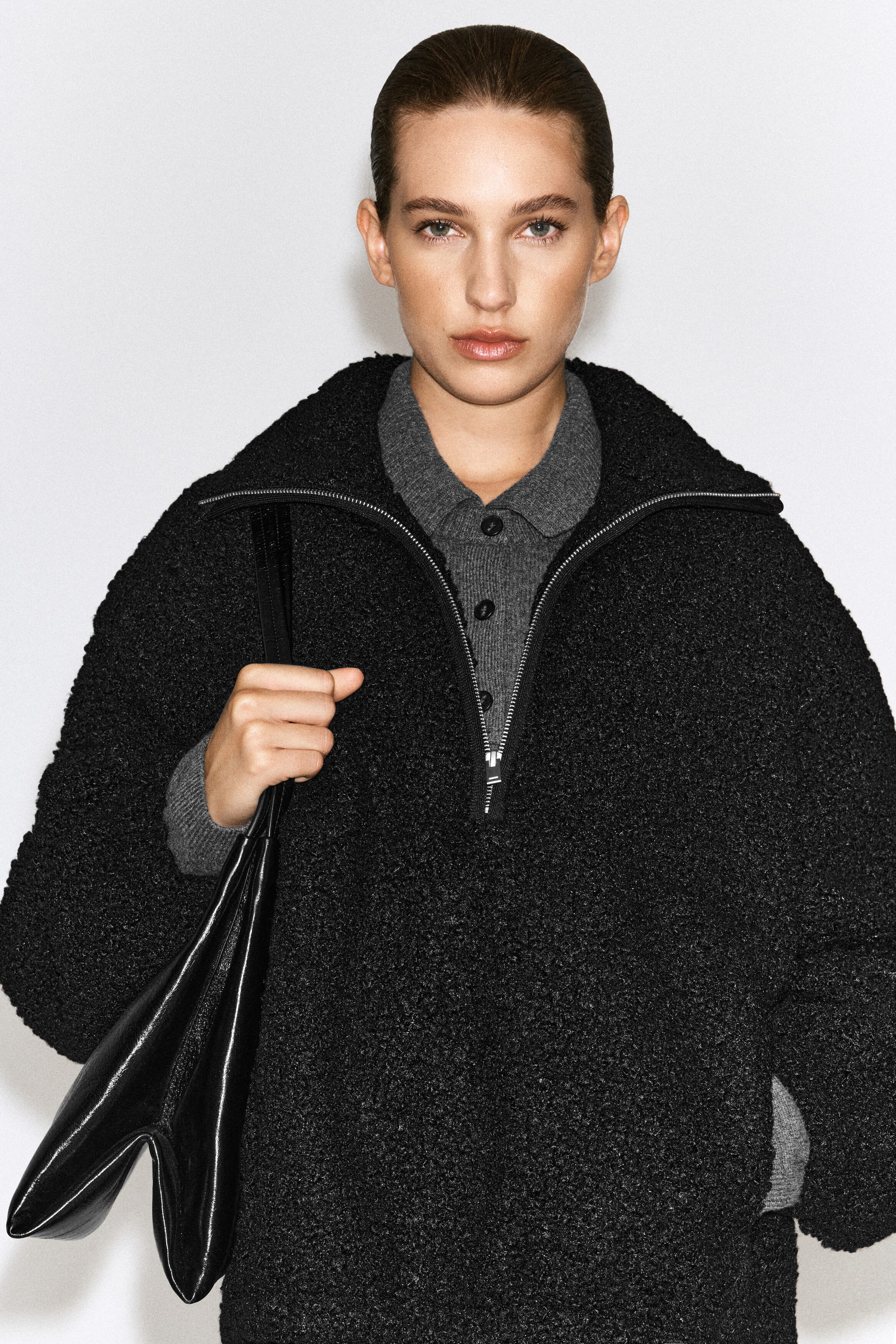 Le chandail zippé en teddy à col montant noir, avec un tissu bouclé texturé et une demi-fermeture éclair argentée, est porté par le modèle par-dessus une chemise en tricot grise boutonnée. Un sac à bandoulière en cuir noir est tenu par sa bandoulière, drapée sur le bras du modèle. Le chandail zippé en teddy à col montant noir, avec un tissu bouclé texturé et une demi-fermeture éclair argentée, est porté par le modèle par-dessus une chemise en tricot grise boutonnée. Un sac à bandoulière en cuir noir est tenu par sa bandoulière, drapée sur le bras du modèle.