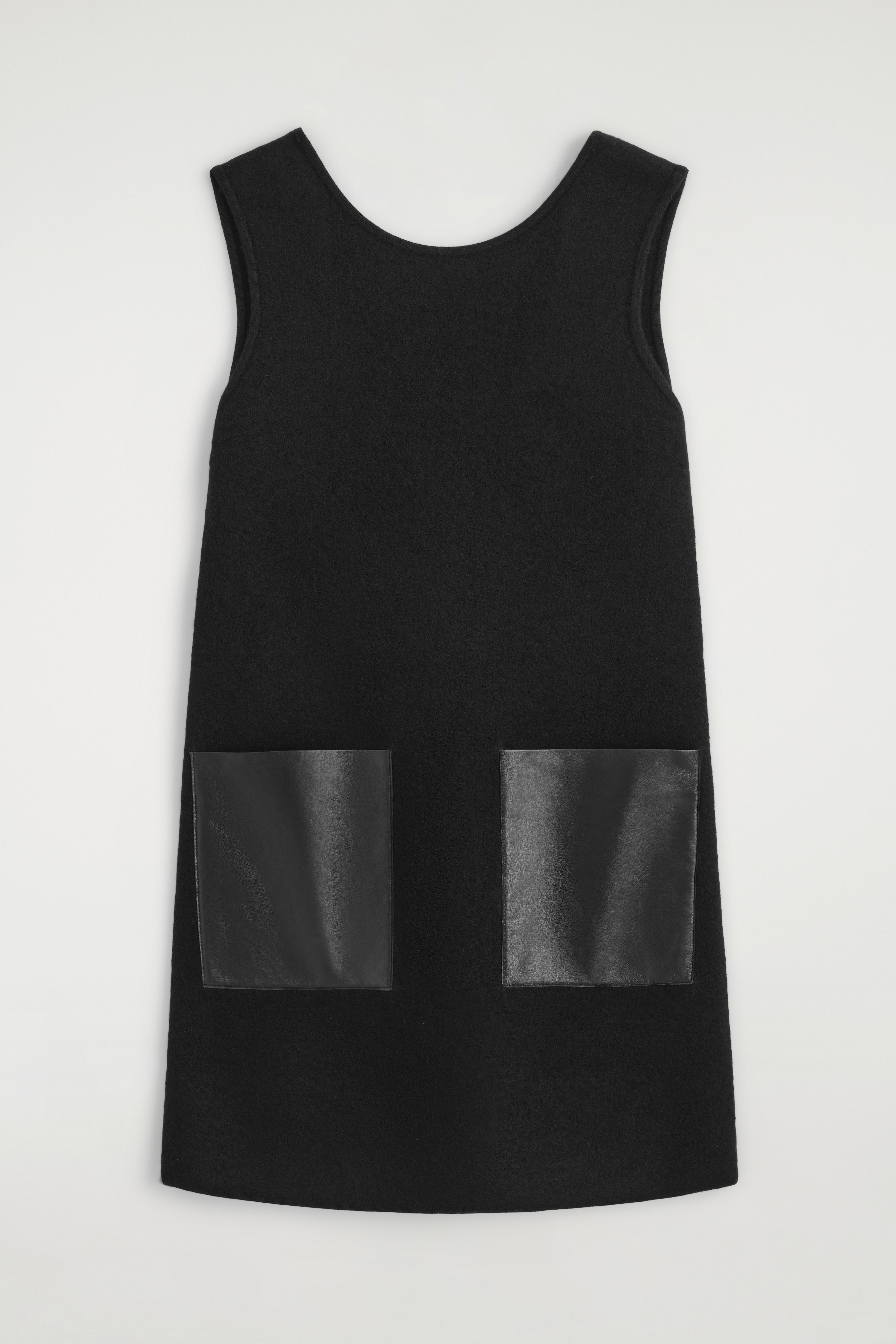 Facing the front, this sleeveless black boiled wool mini dress showcases a shift silhouette. It features a round neckline and two prominent square black leather patch pockets on the front.