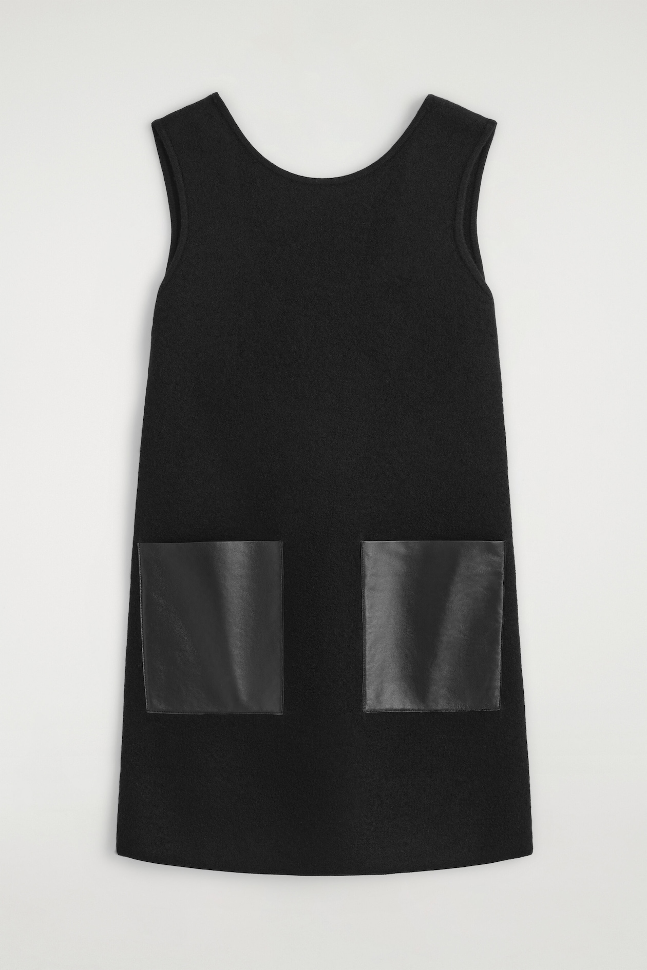 A black sleeveless mini dress is shown from the front, featuring a scoop neckline and a textured boiled wool finish. The dress is designed with a straight silhouette and includes two square black leather patch pockets on the lower front.