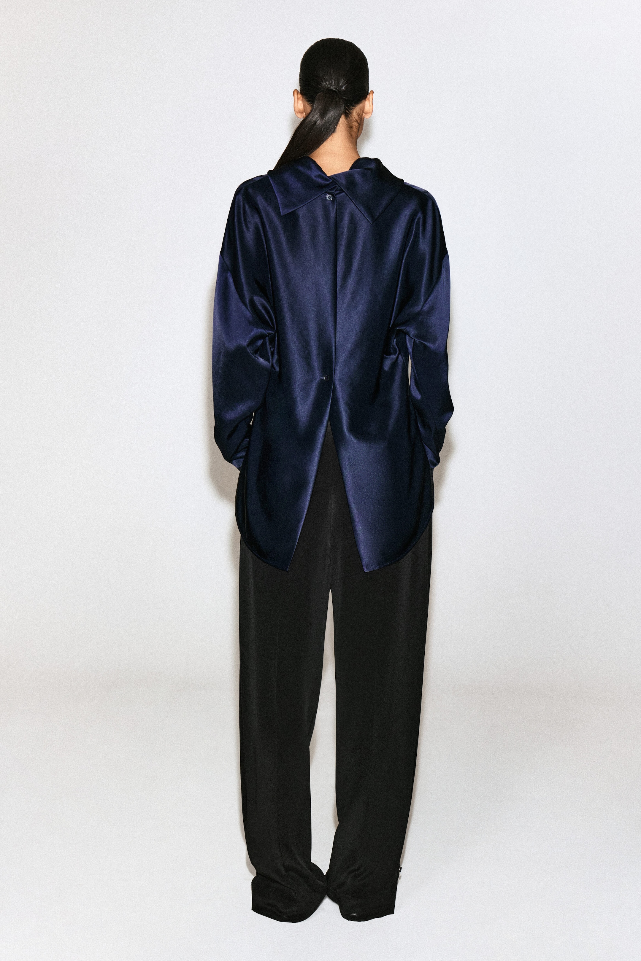 This navy blue satin blouse is shown from the back, highlighting its relaxed fit and lustrous fabric. It features a folded collar detail at the neckline and button closures along a center back seam that opens into a split hem.