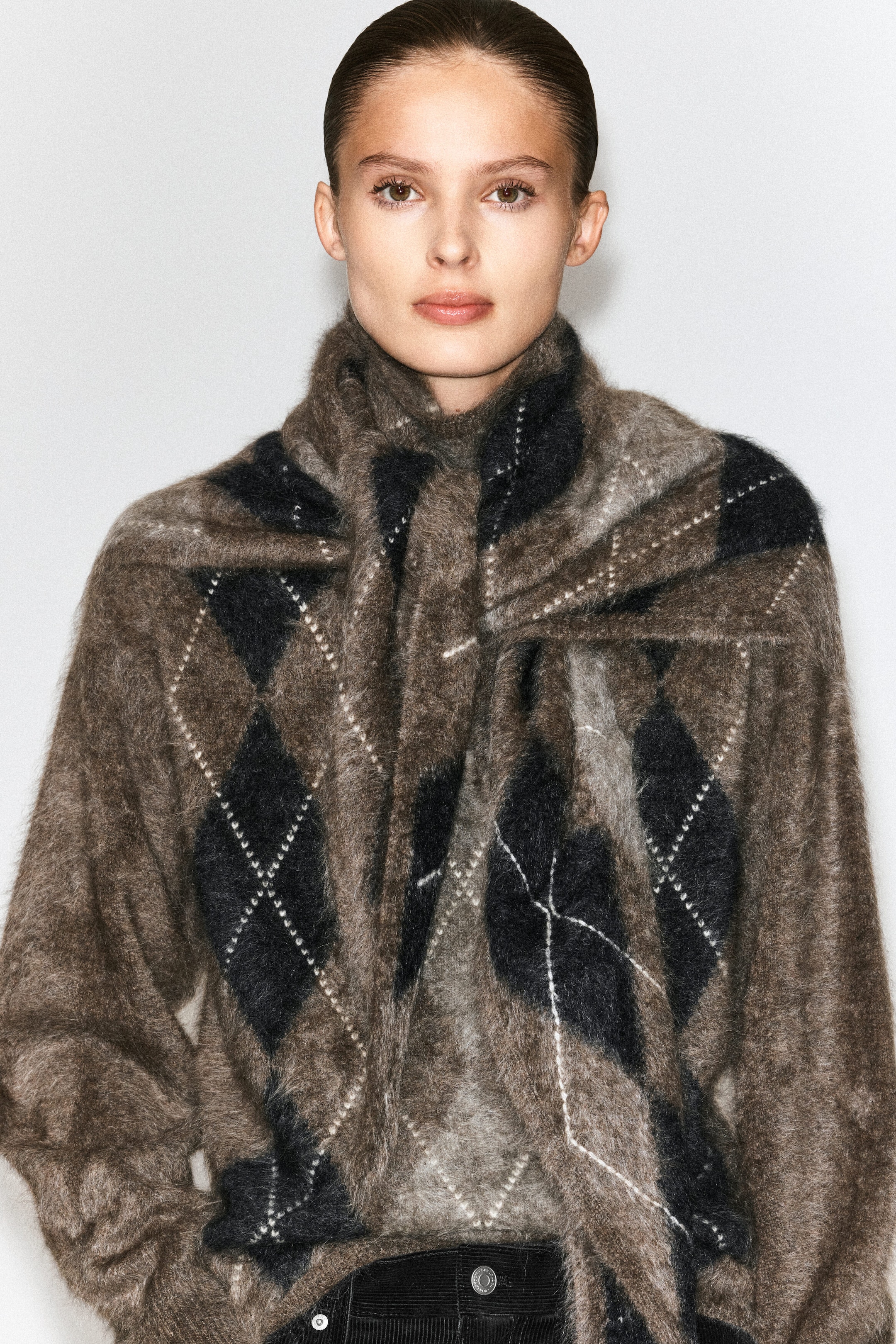 A brushed cashmere argyle triangle scarf in a brown, black, and grey pattern is being worn by the model, wrapped around the neck and draped over the shoulders. The scarf is layered over a matching argyle knit sweater. A brushed cashmere argyle triangle scarf in a brown, black, and grey pattern is being worn by the model, wrapped around the neck and draped over the shoulders. The scarf is layered over a matching argyle knit sweater.