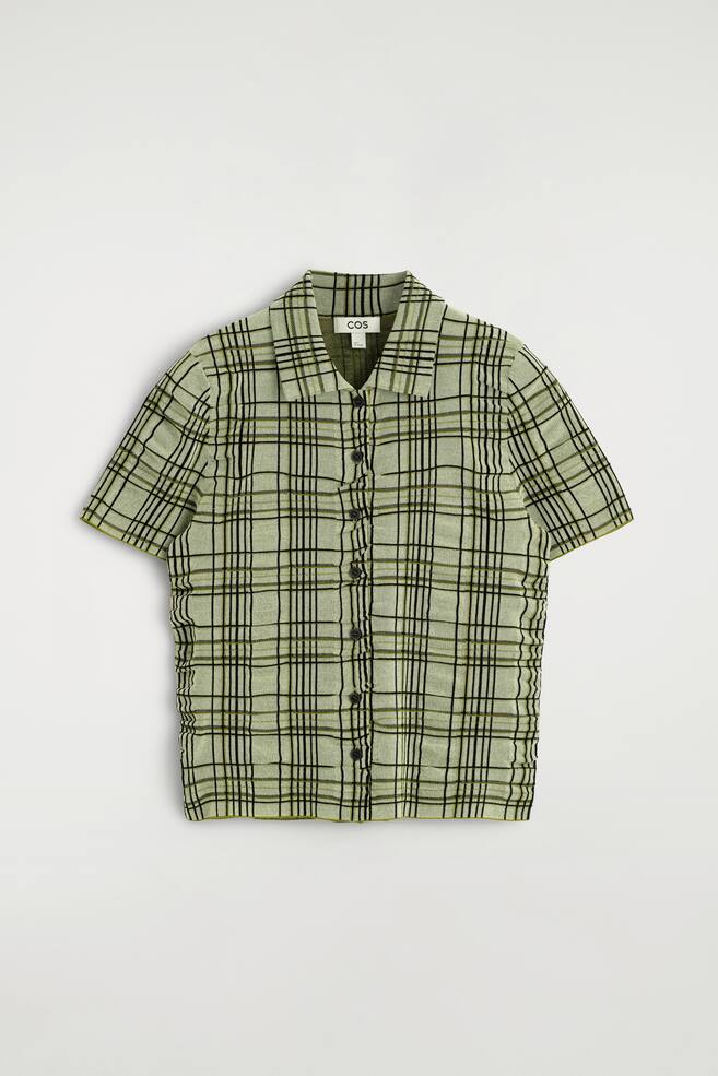 A short-sleeved shirt featuring a light green base with a black and olive green plaid pattern, a point collar, and a full button-front fastening with dark buttons, while displaying a crinkled texture. - 1