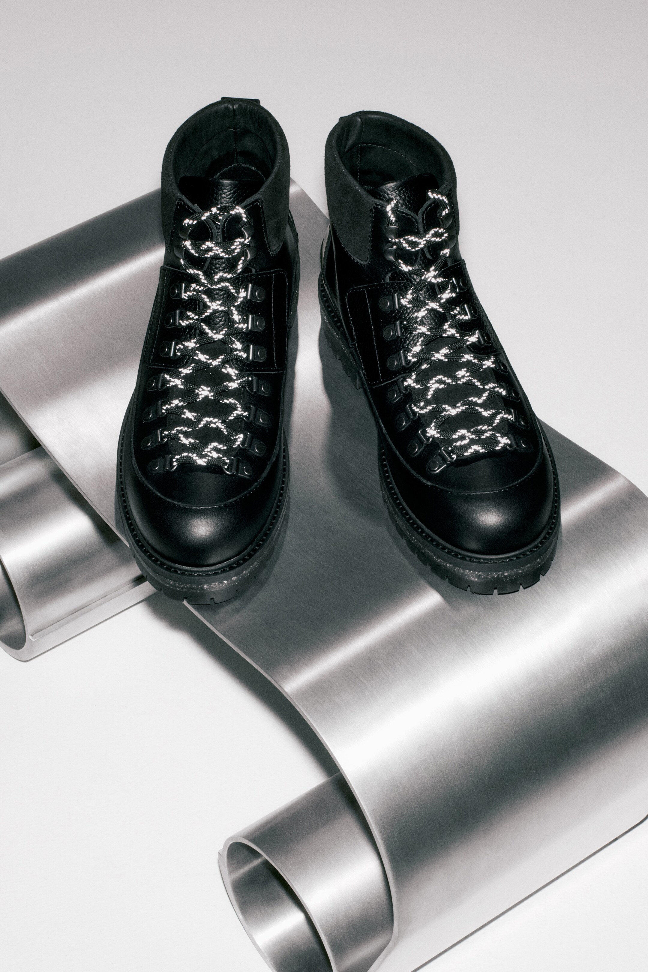 Größeres Bild anzeigen: Ein Paar schwarze, wanderinspirierte Stiefel steht VORNE, gefertigt aus schwarzem Leder mit gepolsterten Wildleder-Knöchelkragen. Die Stiefel verfügen über klobige, geschichtete schwarze Sohlen und schwarze D-Ring-Ösen, durch die weiße und schwarze Seilschnürsenkel mit reflektierenden Details gefädelt sind.