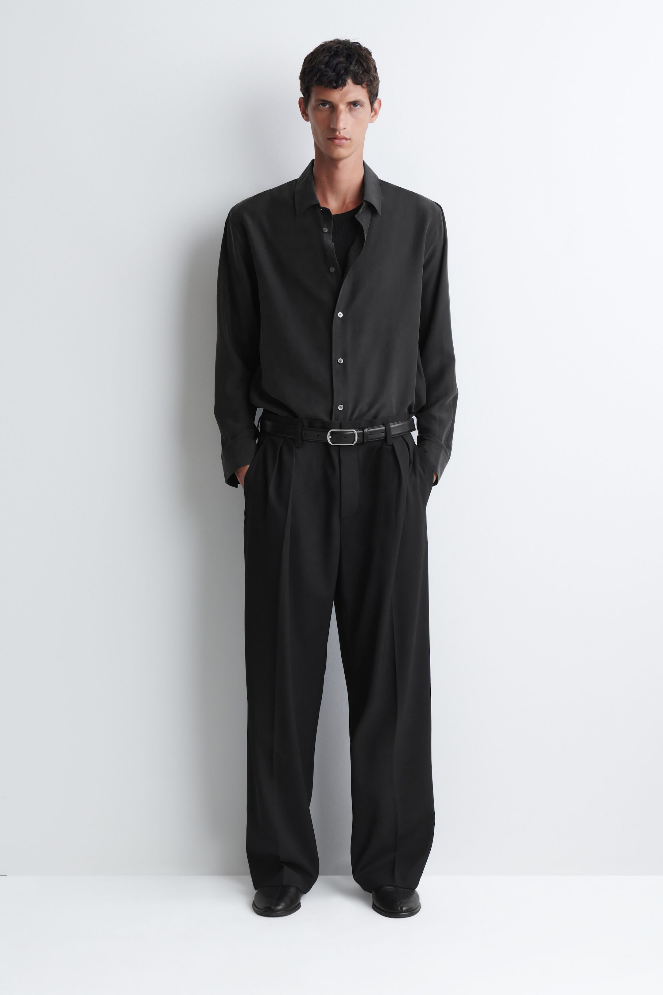 The model is wearing a sandwashed silk shirt in charcoal grey, tucked into wide-leg black trousers with a belt. The relaxed-fit, long-sleeved shirt features a button-up front and a classic collar, layered over a black T-shirt. The model is wearing a sandwashed silk shirt in charcoal grey, tucked into wide-leg black trousers with a belt. The relaxed-fit, long-sleeved shirt features a button-up front and a classic collar, layered over a black T-shirt.
