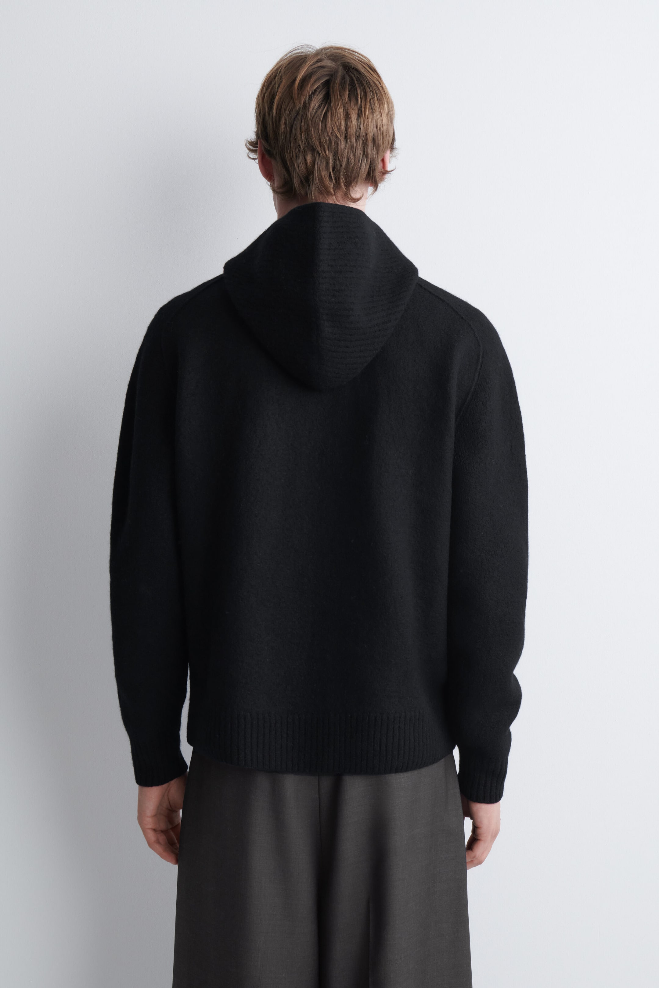 A black merino wool hoodie is being worn by the model, shown from the back. The knitted garment features a relaxed hood resting on the shoulders, ribbed cuffs, and a ribbed hem, and is styled with dark grey trousers. A black merino wool hoodie is being worn by the model, shown from the back. The knitted garment features a relaxed hood resting on the shoulders, ribbed cuffs, and a ribbed hem, and is styled with dark grey trousers.